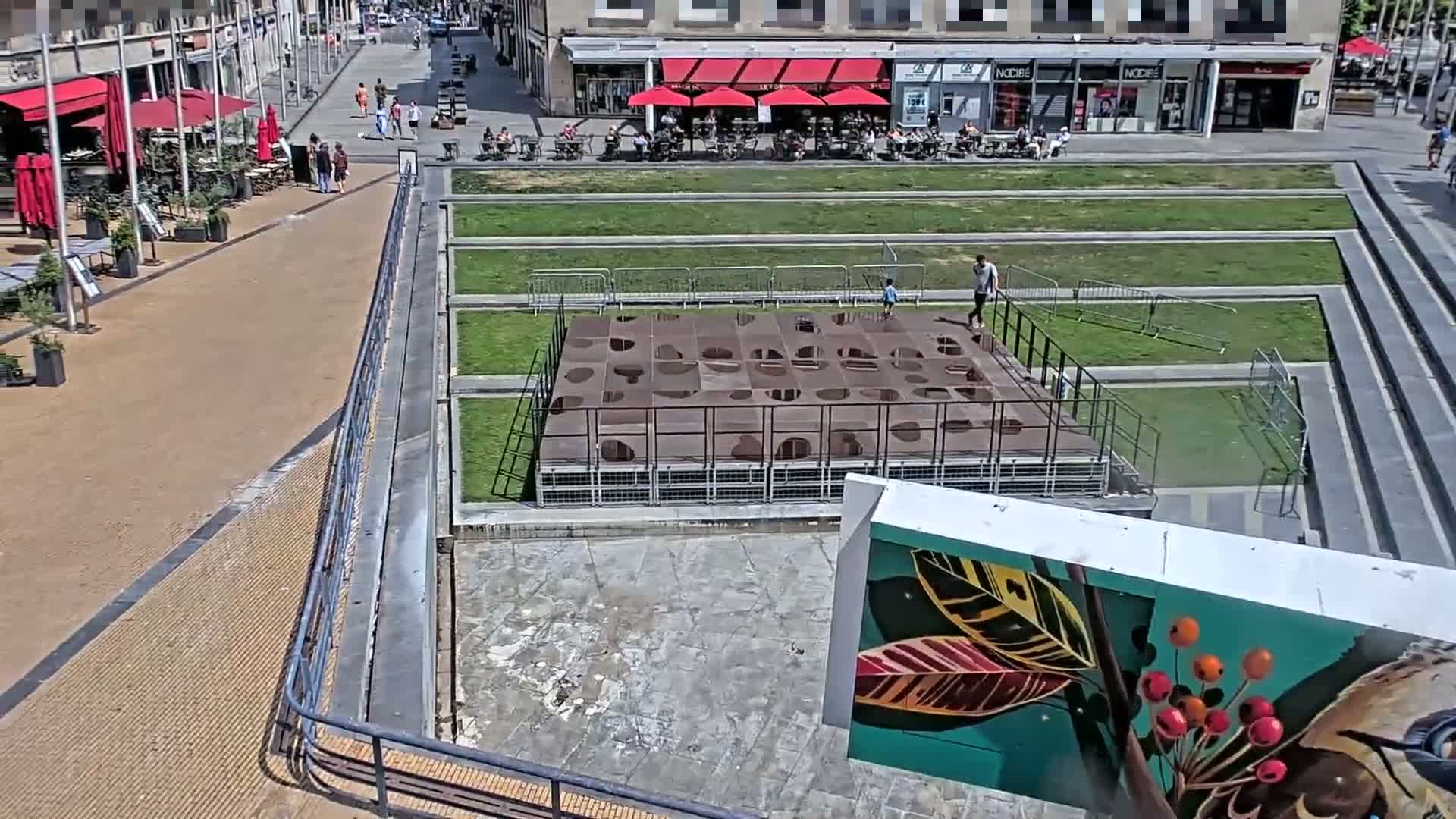 A high-angle view shows a city square on a sunny day, featuring a brown gridded structure, grassy areas, and a mural of colorful leaves and berries.