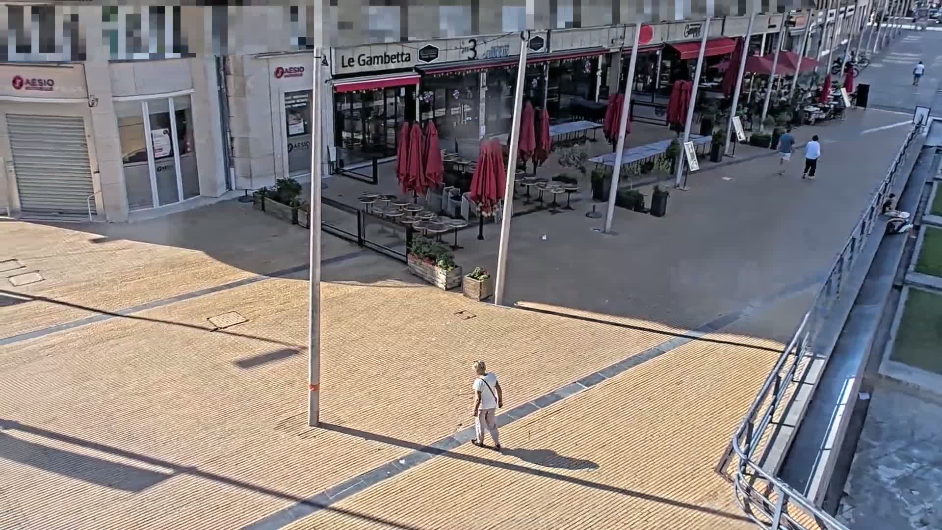 A person walks across a sun-drenched paved plaza with an outdoor cafe featuring red umbrellas and empty tables.