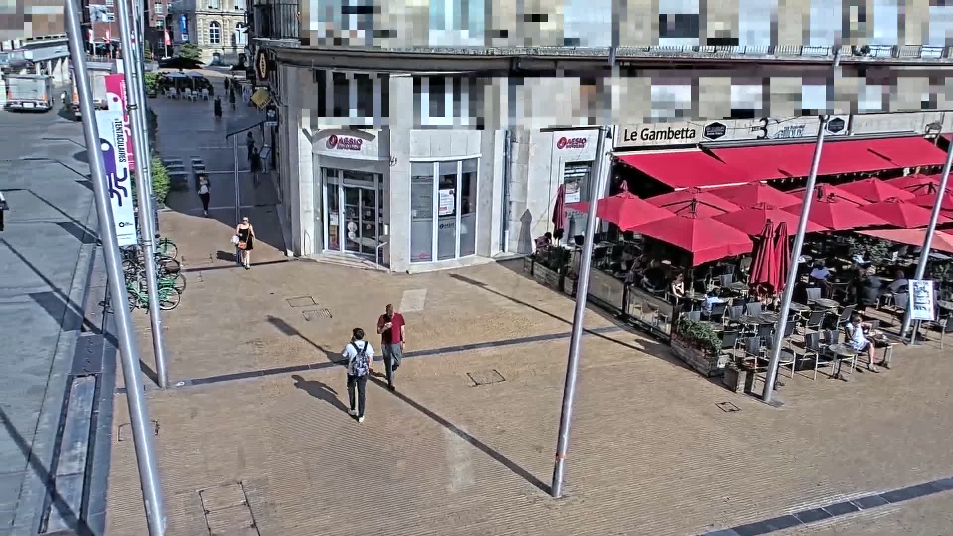 A sunny day shows a paved plaza with pedestrians, bicycles, and an outdoor cafe with red umbrellas.