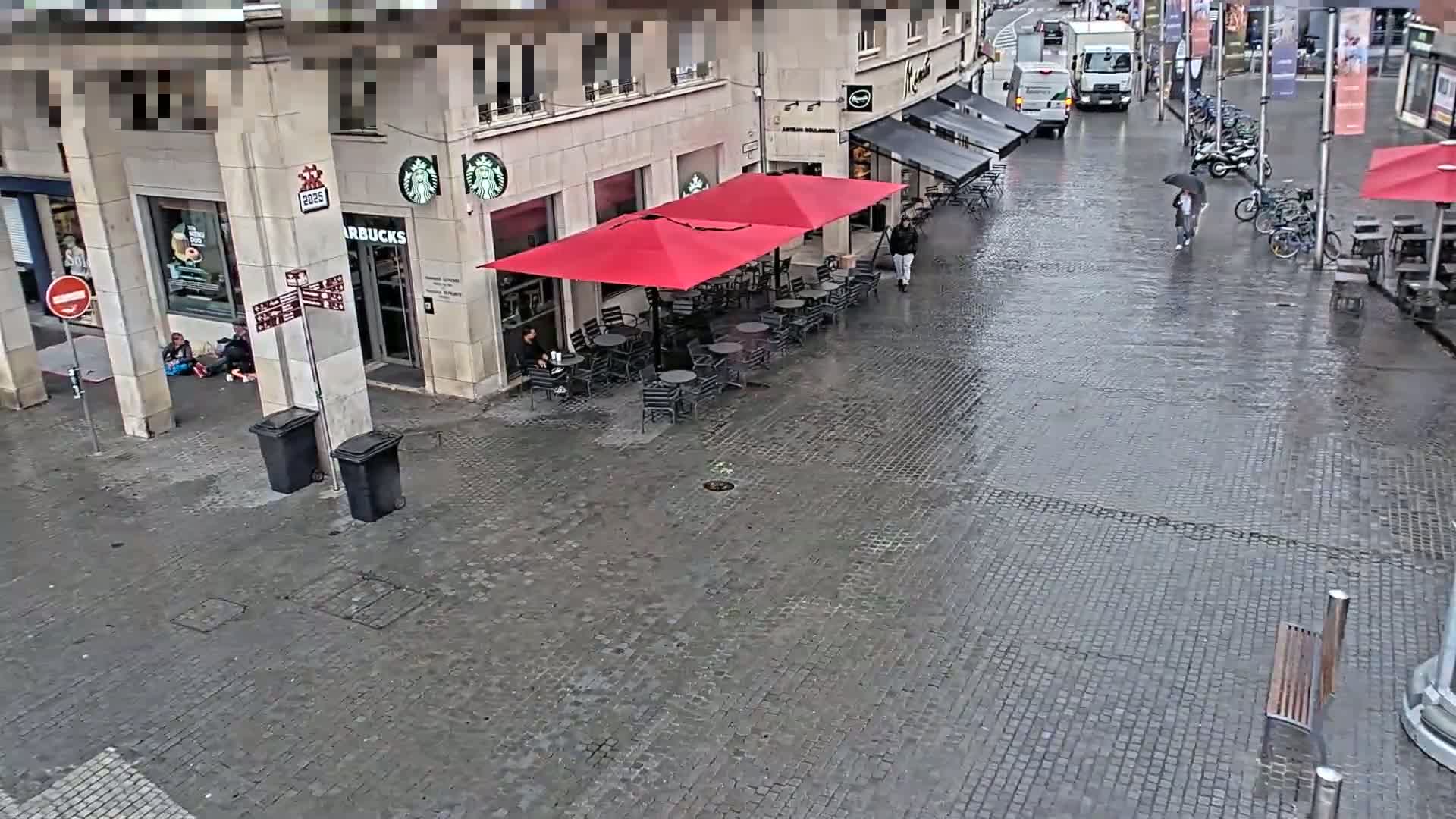 A wet cobblestone street shows a Starbucks with outdoor seating under red umbrellas, a few pedestrians, and parked bicycles.