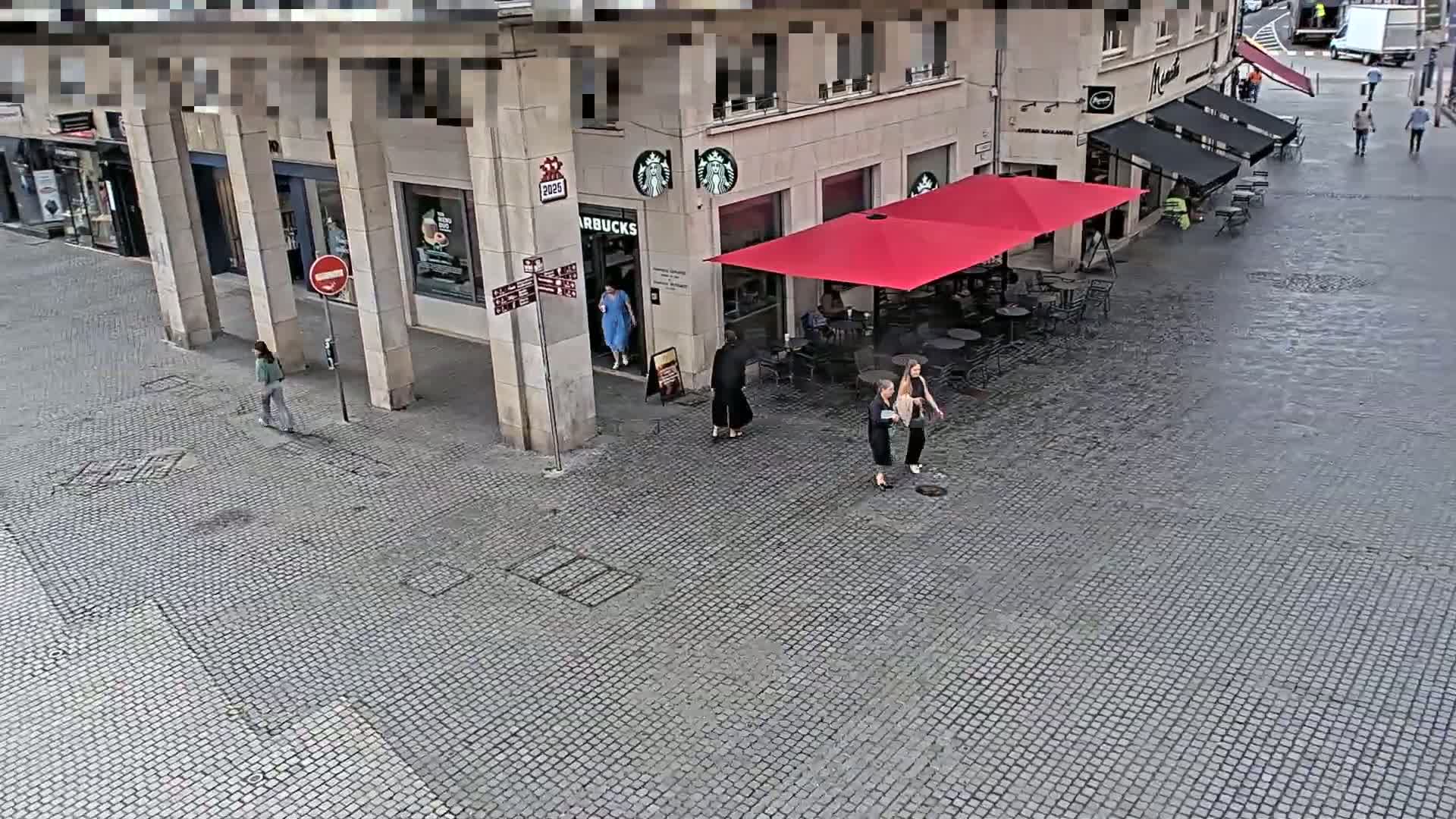 A cobblestone street scene shows a Starbucks with a red patio umbrella and several pedestrians, under a mostly sunny sky.