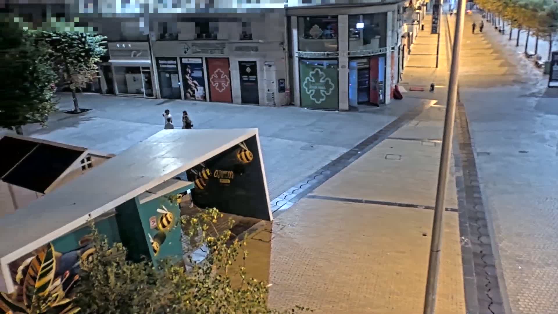 At night, a mostly empty plaza and street scene features a few pedestrians, buildings with shops on the ground floor, and a structure with a bee motif.