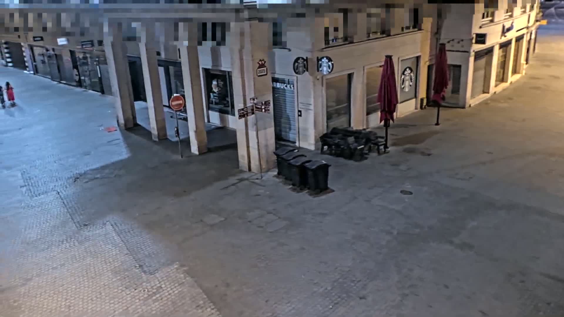 A mostly empty street at night, with a Starbucks visible, shows a few people in the distance and closed-up outdoor seating.