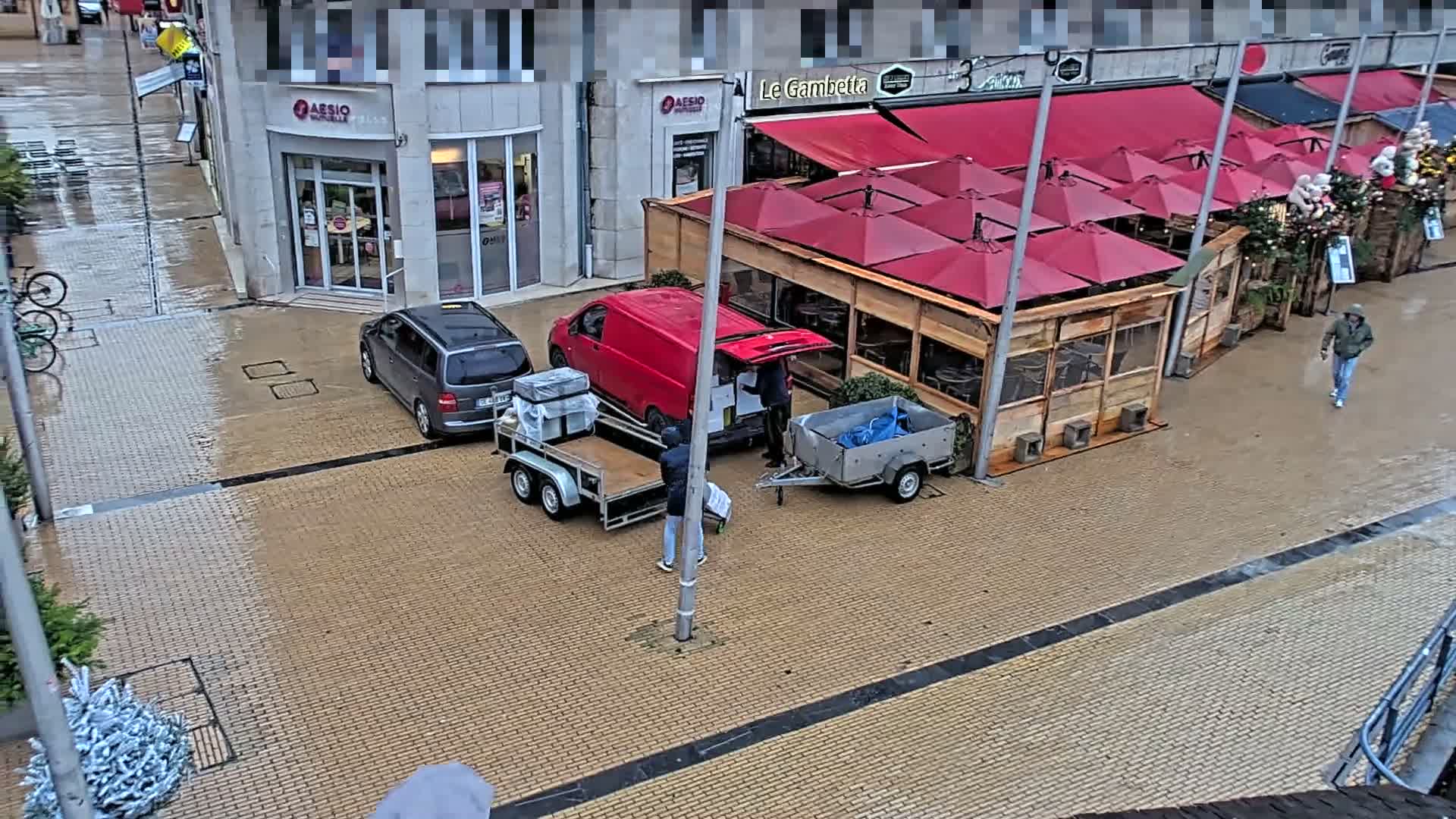 On a wet and overcast day, a street lined with buildings and a row of red-awning-covered wooden market stalls shows a red van and trailers being unloaded, a parked minivan, and pedestrians on the damp brick pavement.