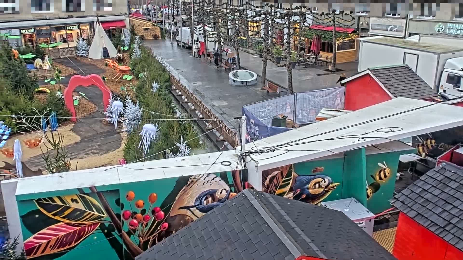 An elevated view captures a festive outdoor display featuring a red heart arch, evergreen trees, and whimsical jellyfish decorations adjacent to a wet paved urban street lined with bare trees, pedestrians, and various temporary market stalls, under damp conditions.