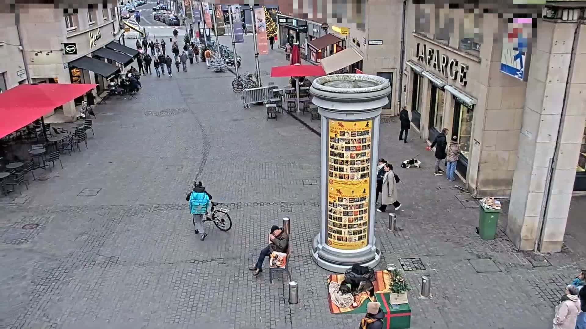 An elevated view captures a festive outdoor display featuring a red heart arch, evergreen trees, and whimsical jellyfish decorations adjacent to a wet paved urban street lined with bare trees, pedestrians, and various temporary market stalls, under damp conditions.