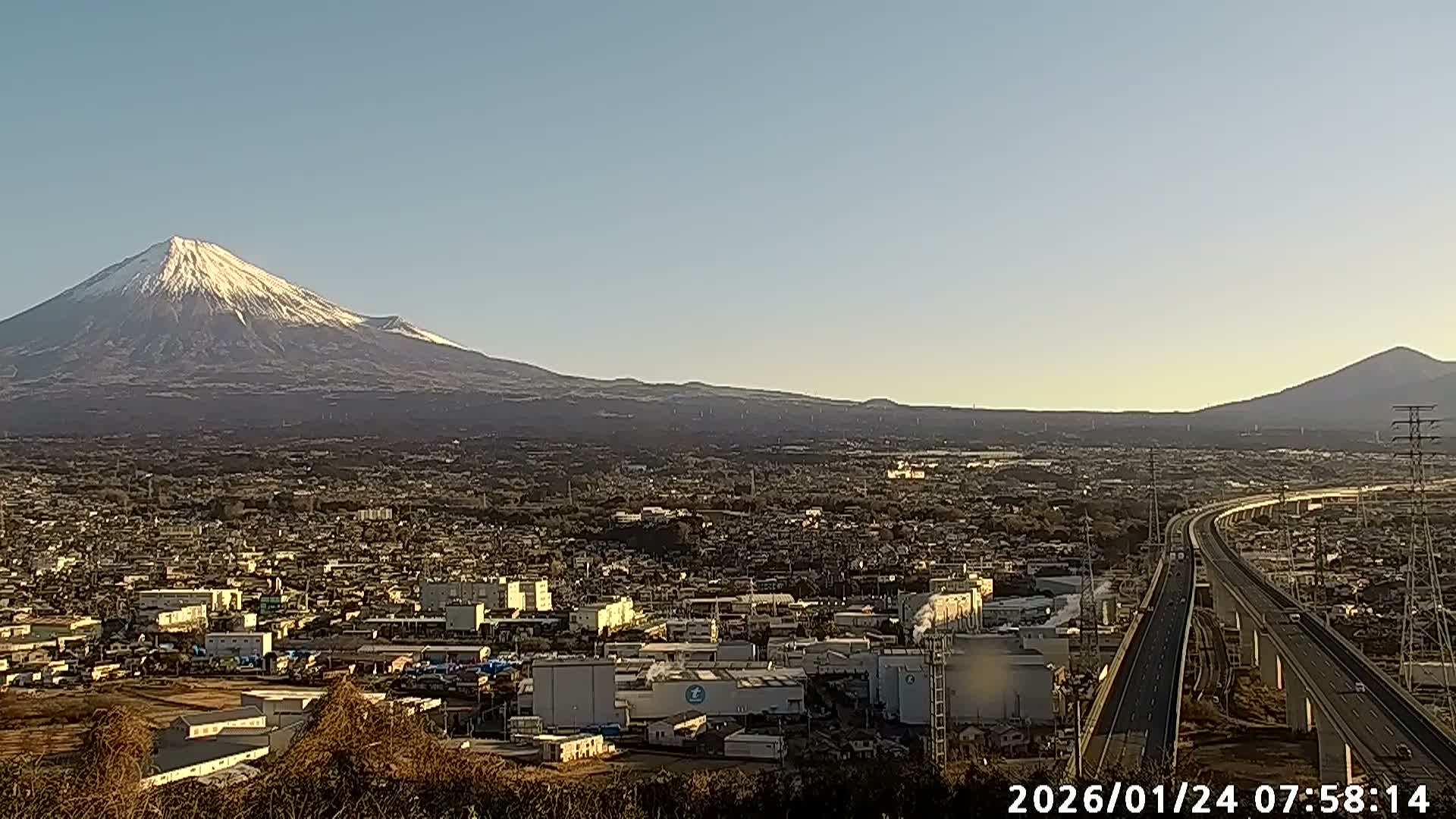 A wide shot captures the snow-capped Mount Fuji towering over a sprawling city with an elevated highway, all bathed in the warm light of a clear, early morning sky.