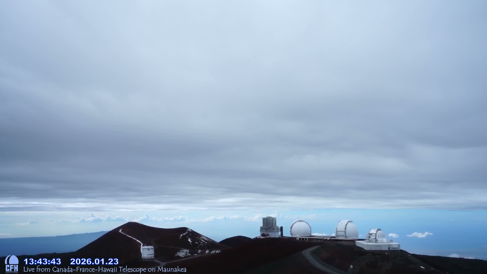 A wide outdoor view shows several white observatory domes and structures atop dark, reddish-brown mountain peaks with a winding road, all beneath a heavily overcast grey sky, while a clear blue expanse of ocean or lower clouds is visible far below the peaks.