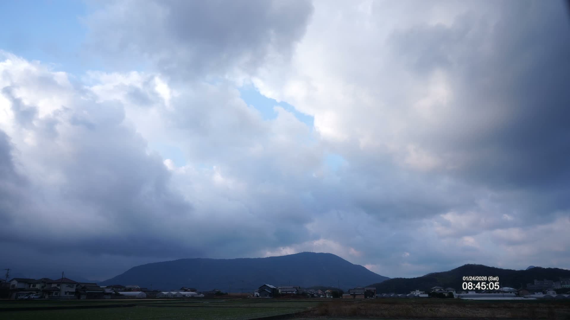 Under a mostly cloudy sky filled with heavy grey clouds and glimpses of blue, a rural landscape unfolds with agricultural fields, scattered houses, and a prominent dark mountain range on the horizon.