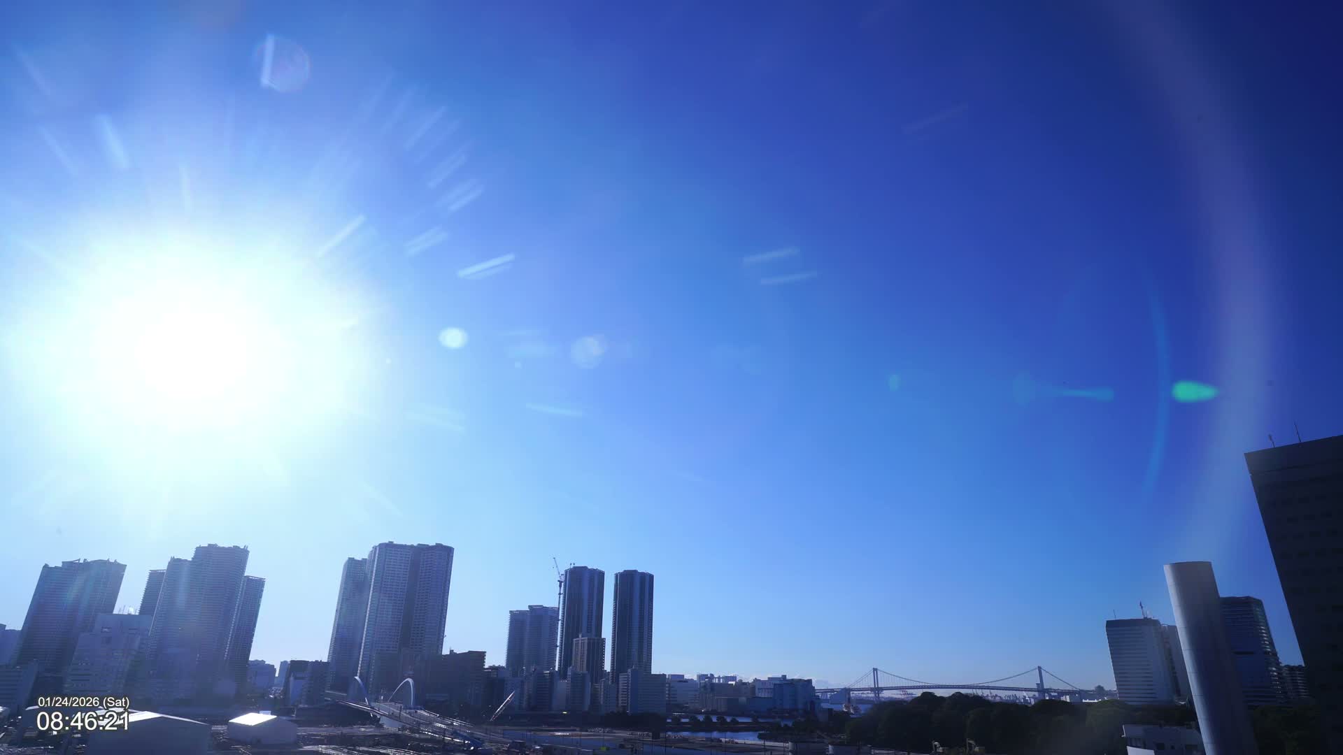 An urban skyline featuring numerous high-rise buildings, a body of water, and a prominent suspension bridge in the distance is visible under a brilliant, clear blue sky with strong sunlight and lens flare.