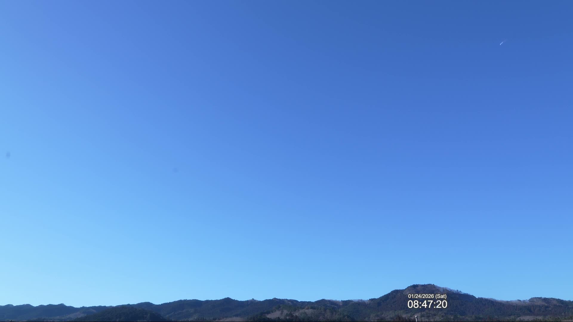 A wide shot captures a vast, clear blue sky with a faint contrail, stretching above a distant range of dark, tree-covered mountains on a clear day.