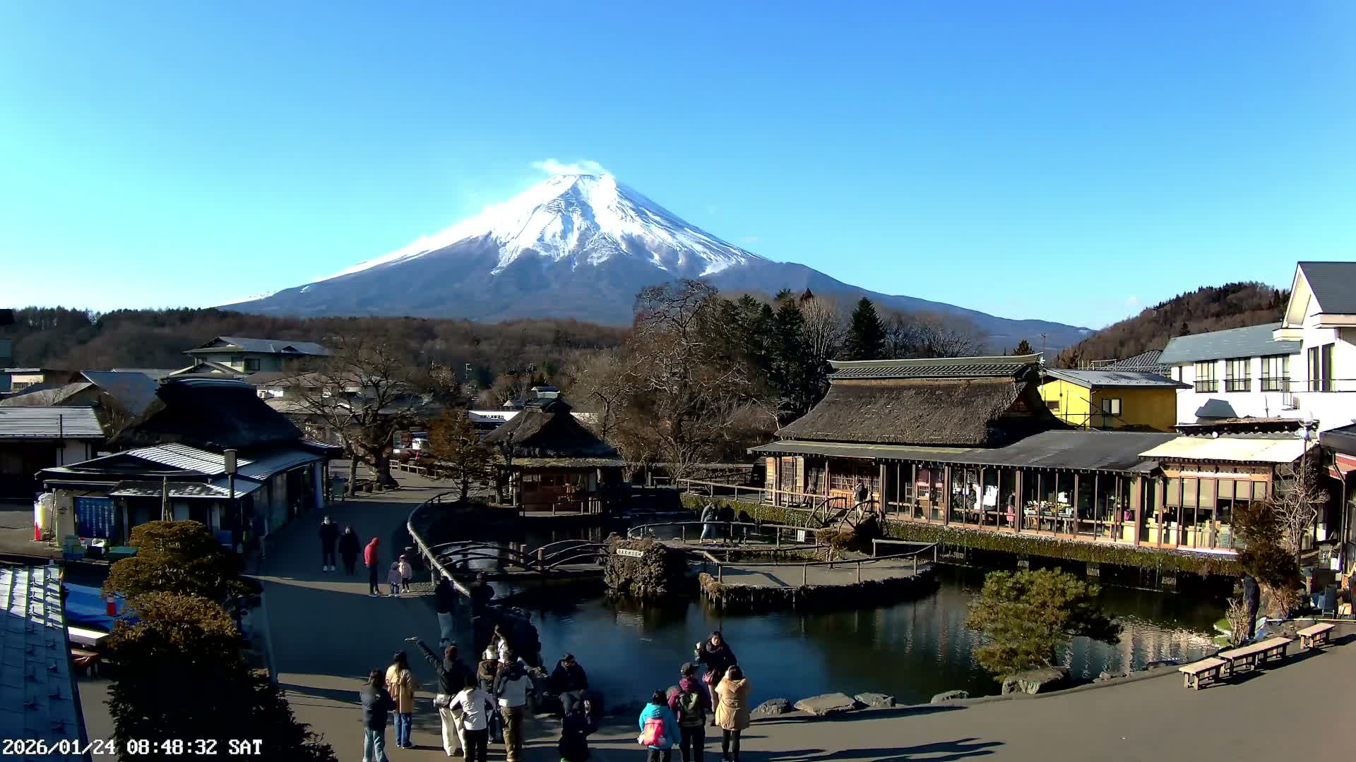 The image displays a bustling traditional Japanese village with thatched-roof buildings and a serene pond under a clear blue, sunny sky, with the majestic, snow-capped Mount Fuji prominent in the background.