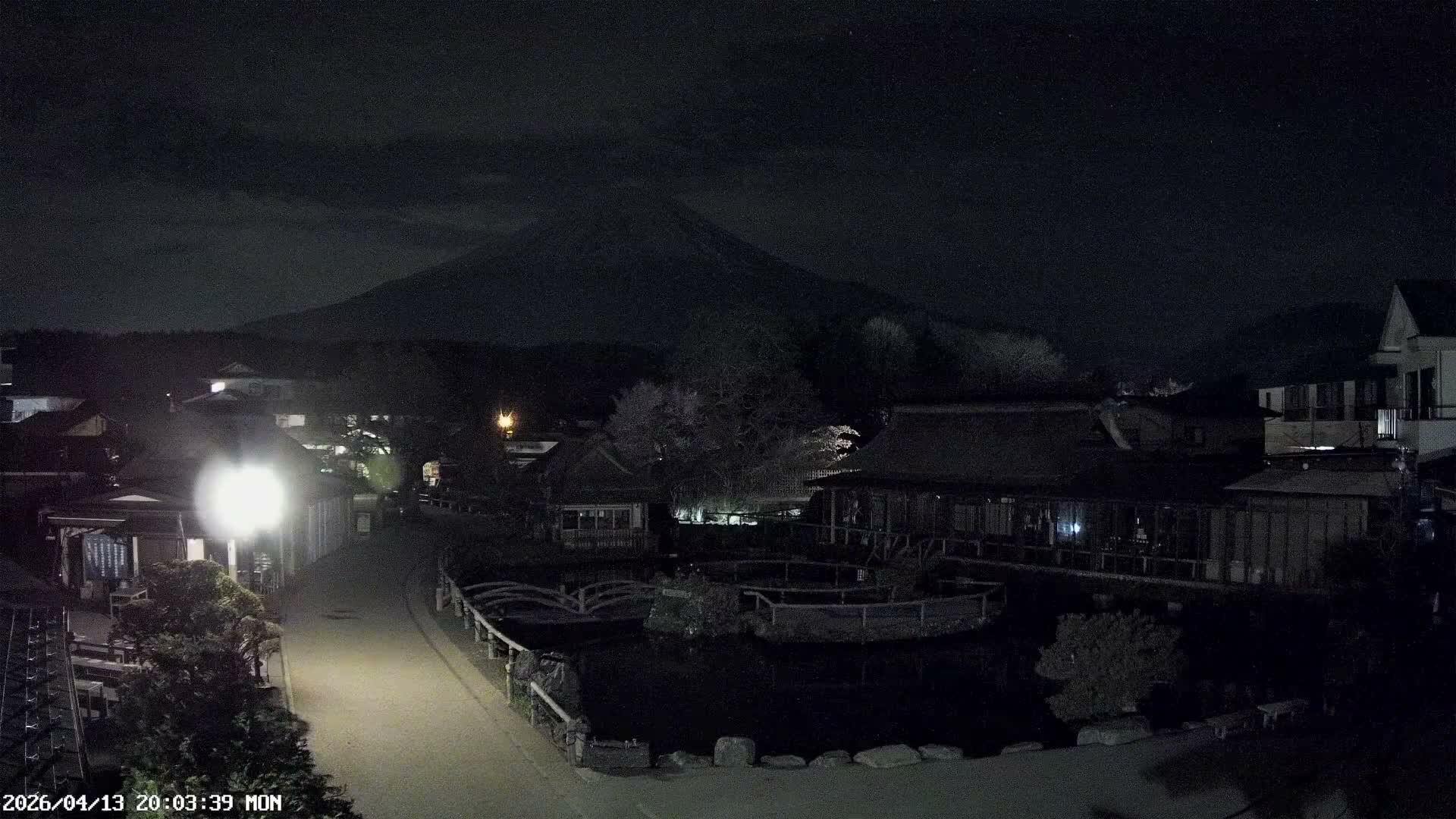 The image displays a bustling traditional Japanese village with thatched-roof buildings and a serene pond under a clear blue, sunny sky, with the majestic, snow-capped Mount Fuji prominent in the background.