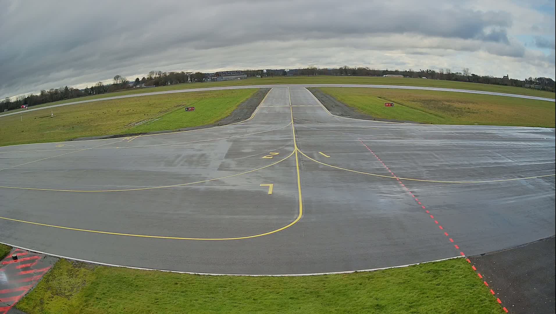 A wet airport tarmac with yellow and red lines indicating taxiways and runways under a cloudy sky.