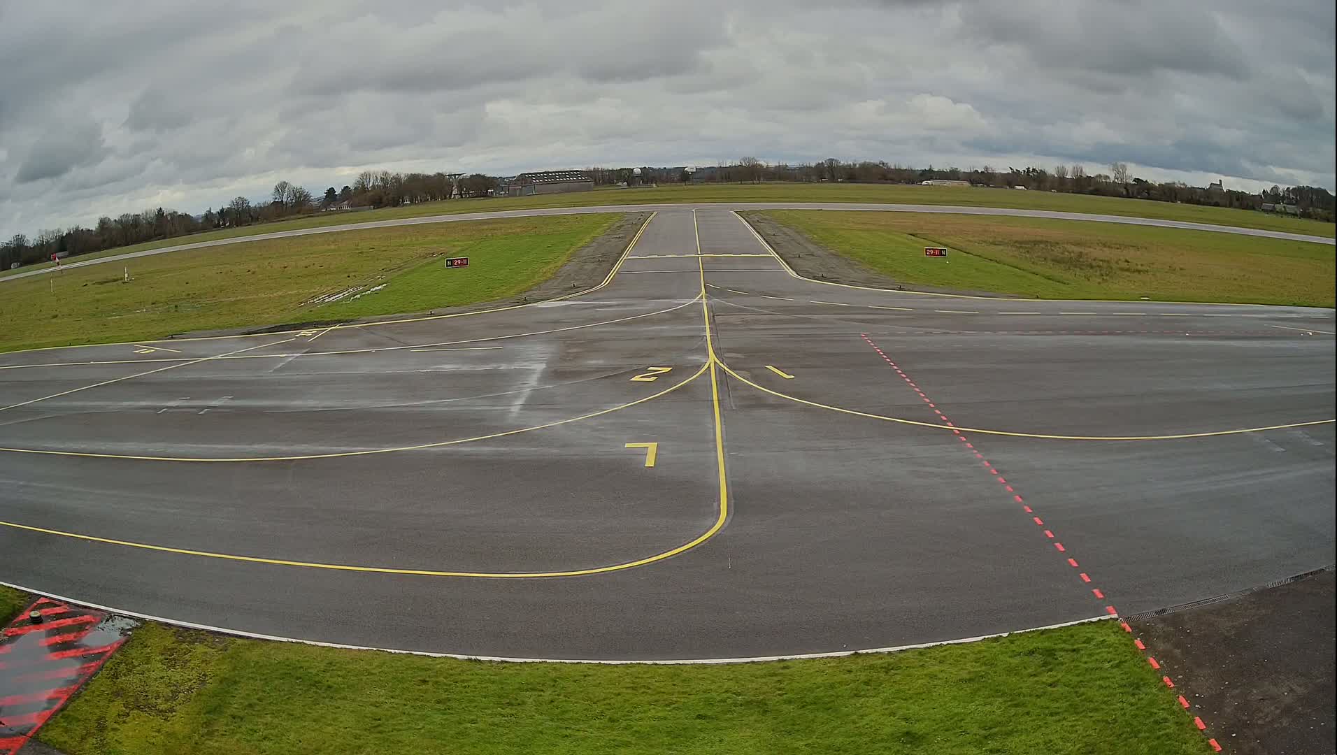 A wet airport runway with yellow and red markings is seen under a cloudy sky.