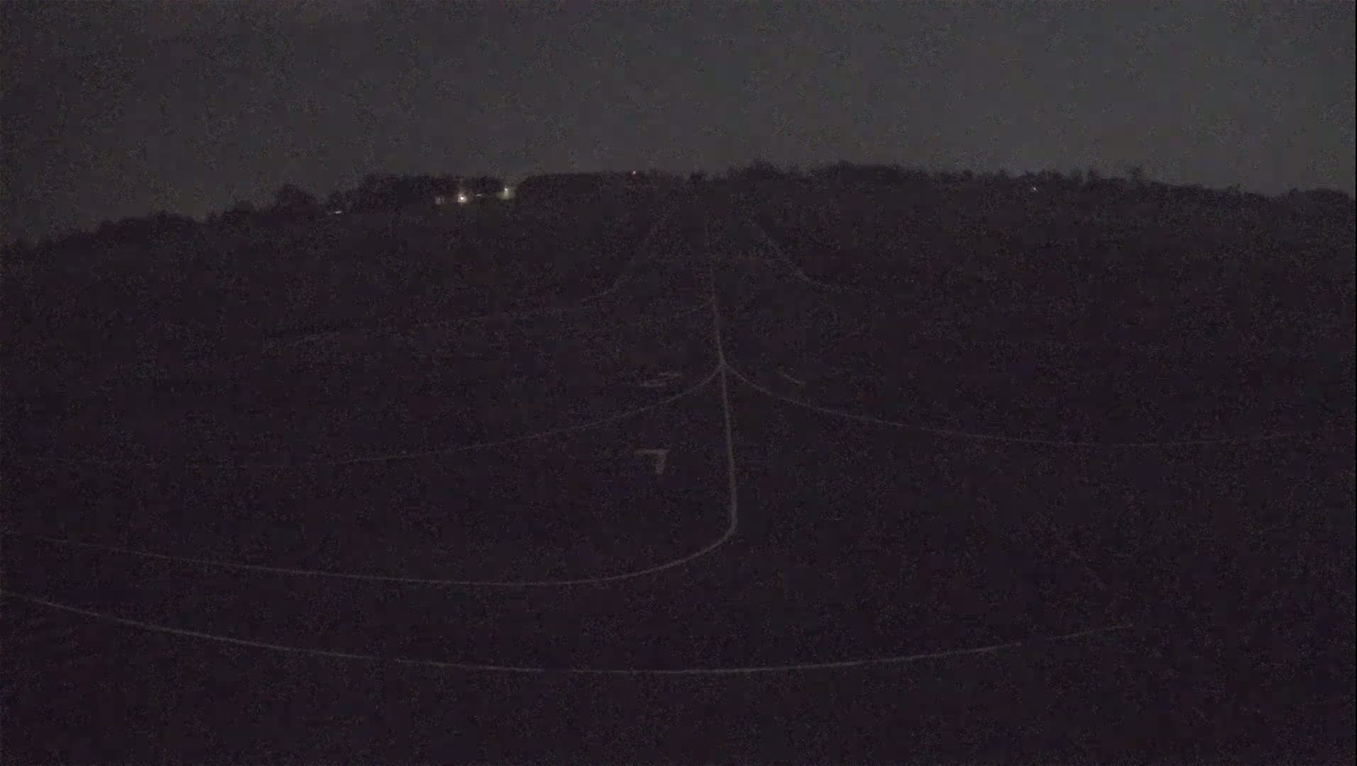 A dark, nighttime view of a field with faintly visible lines and a distant, dimly lit hill in the background.