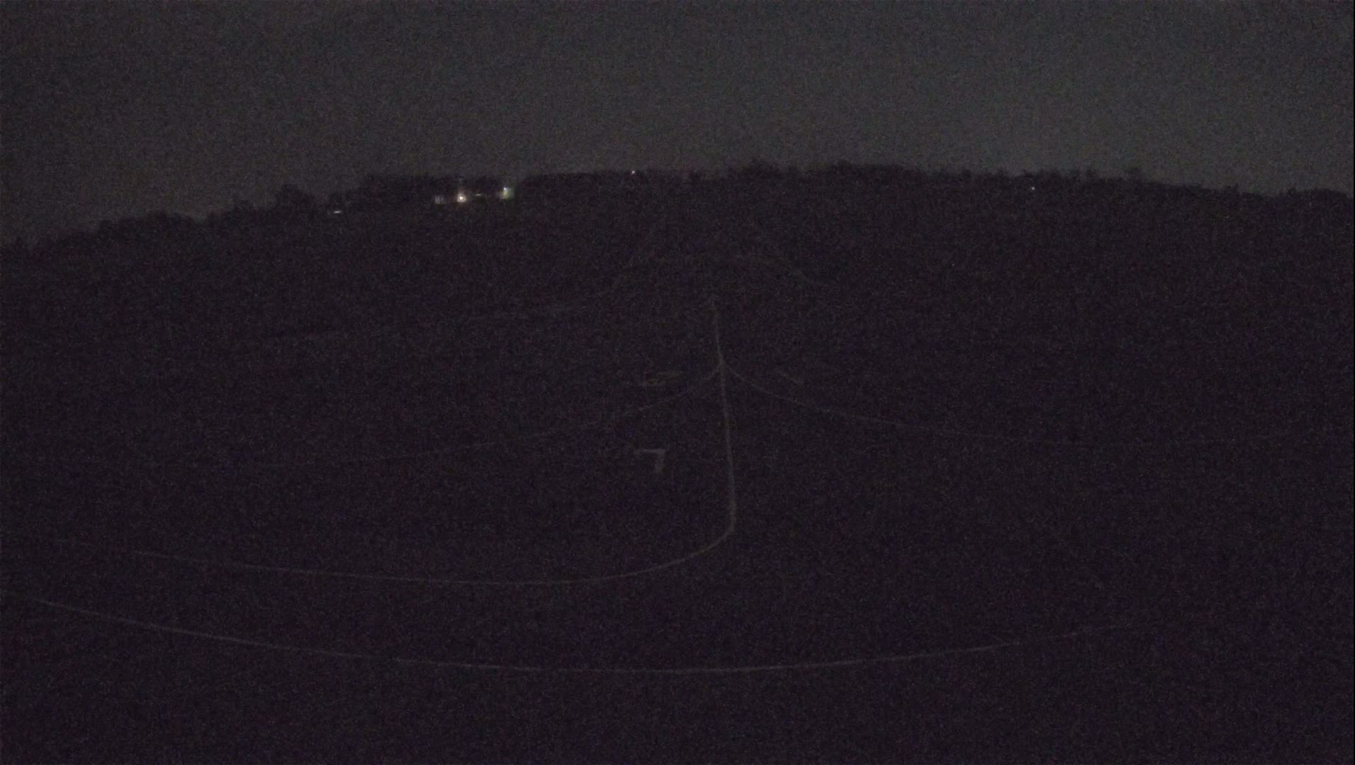 A dark, nighttime view of a mostly flat, dark landscape with faint lines and a few distant lights.