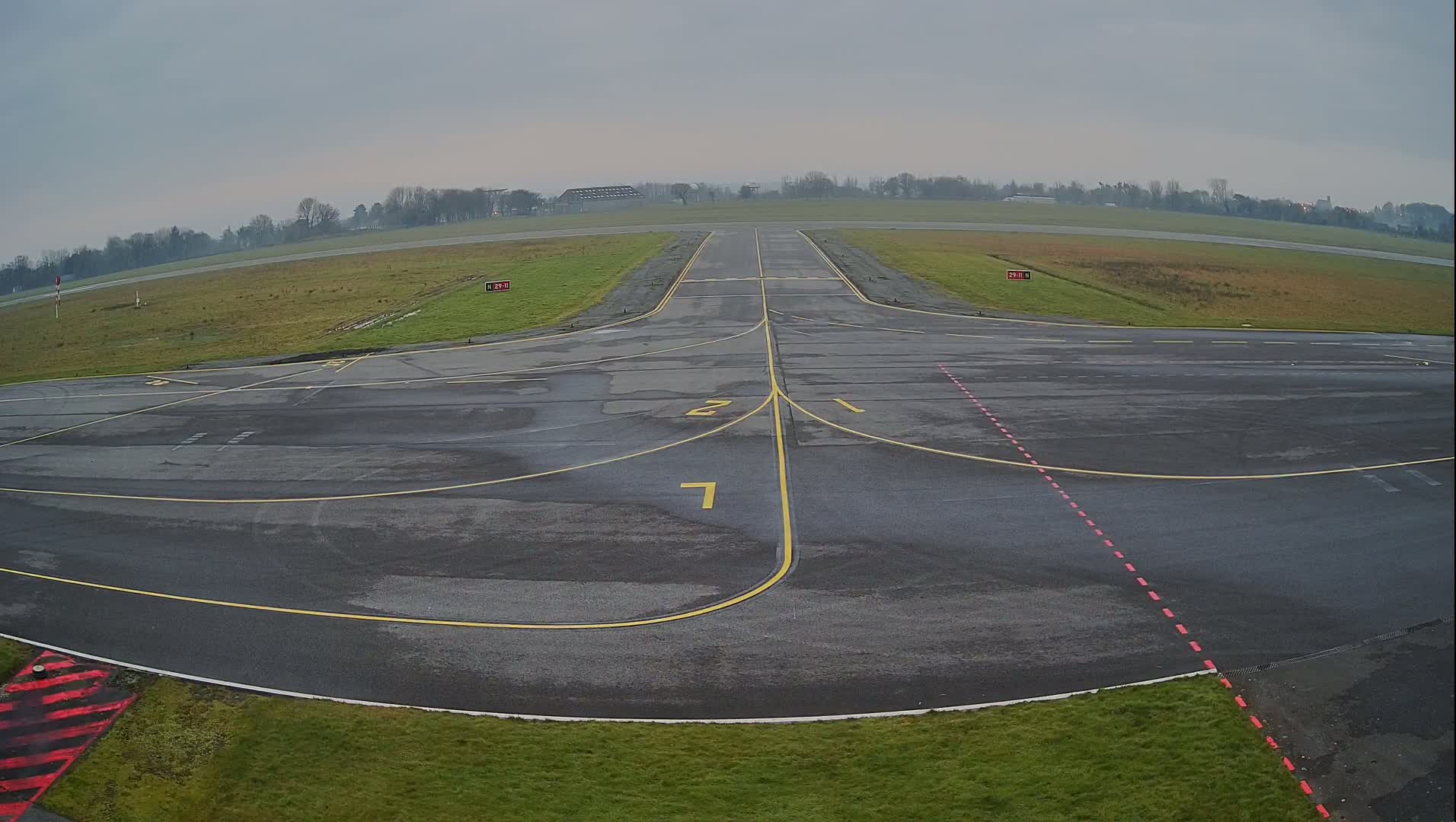 A wet, gray airfield with yellow and red markings on the tarmac extends to a grassy runway under an overcast sky.