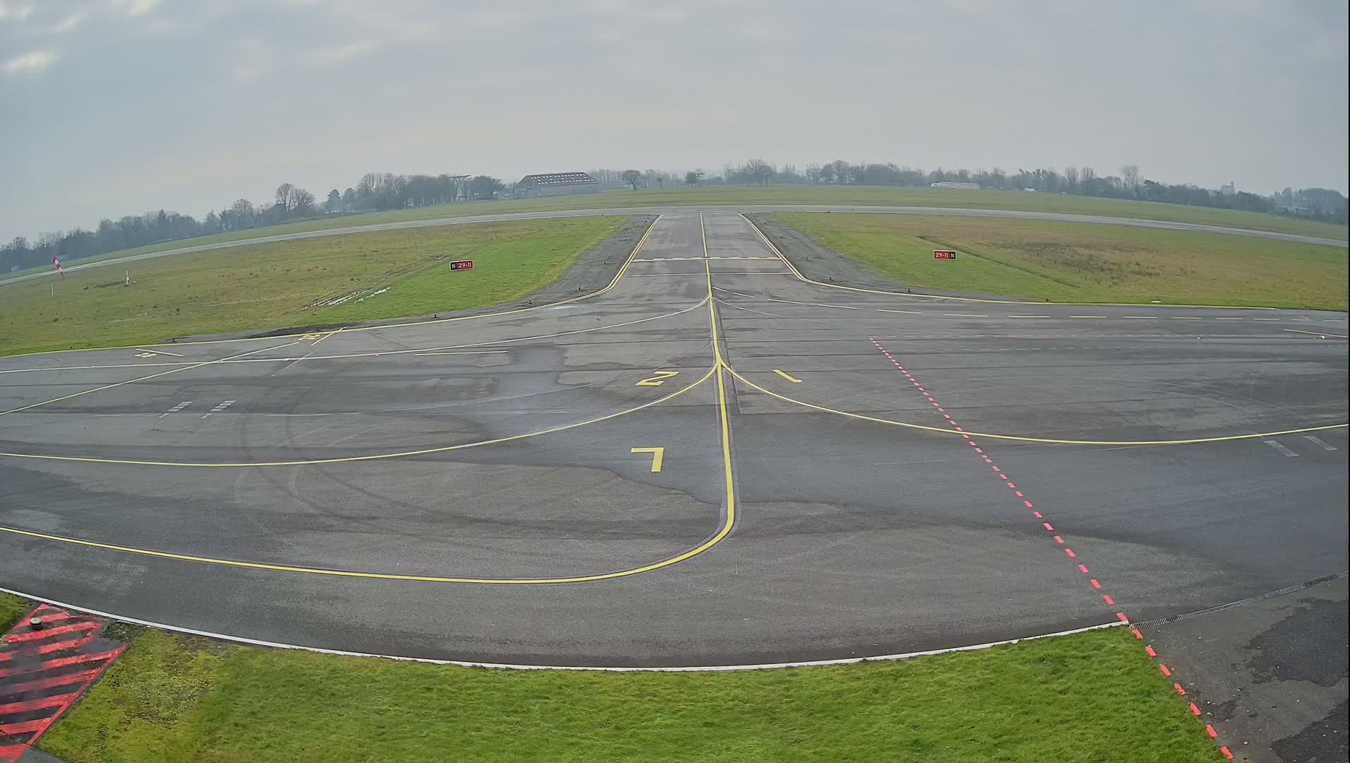 A wet airport tarmac with yellow and red markings, under an overcast sky.