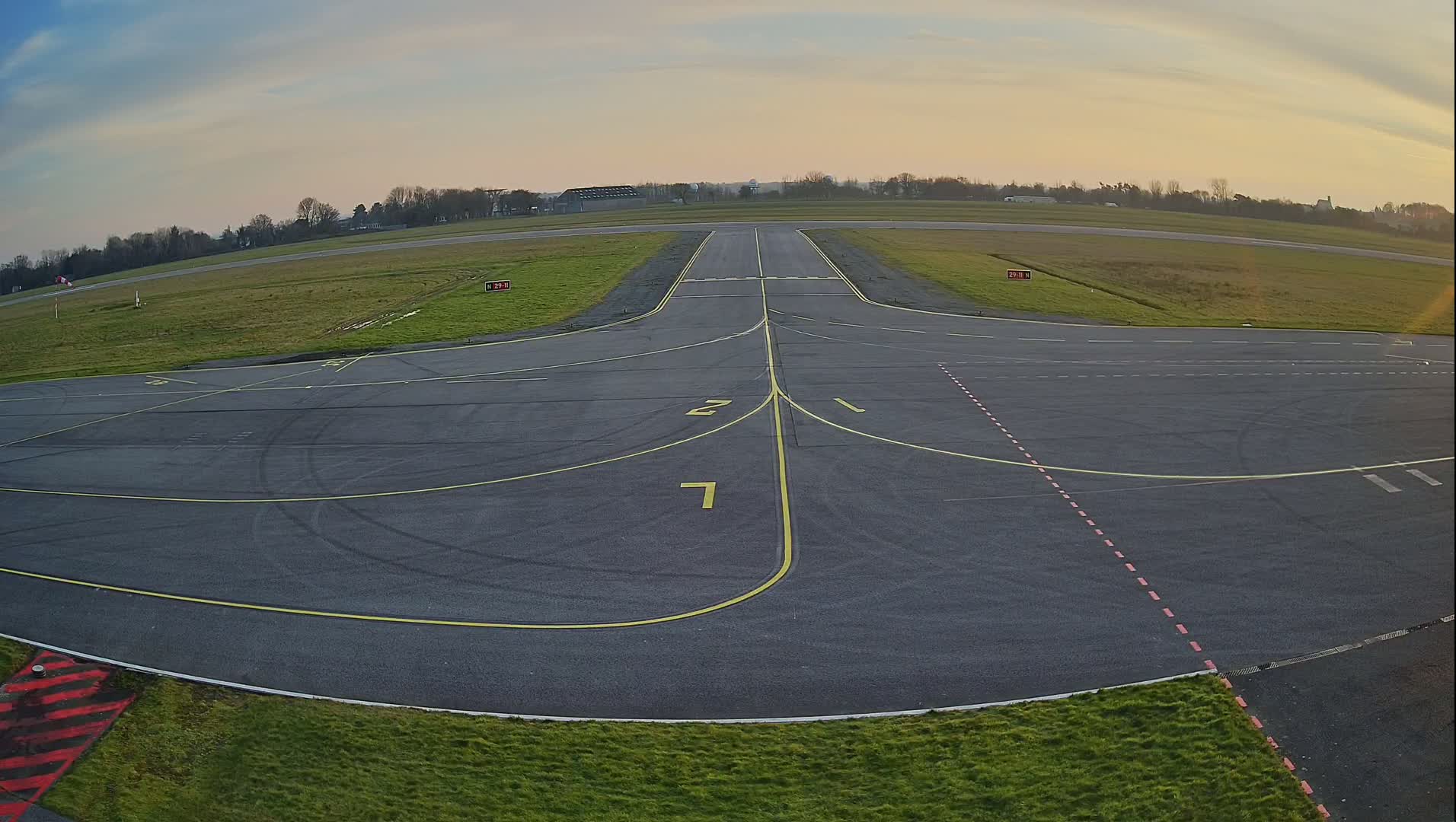 A tarmac airport runway and taxiway intersect, surrounded by short green grass under a partly cloudy sky at sunset.