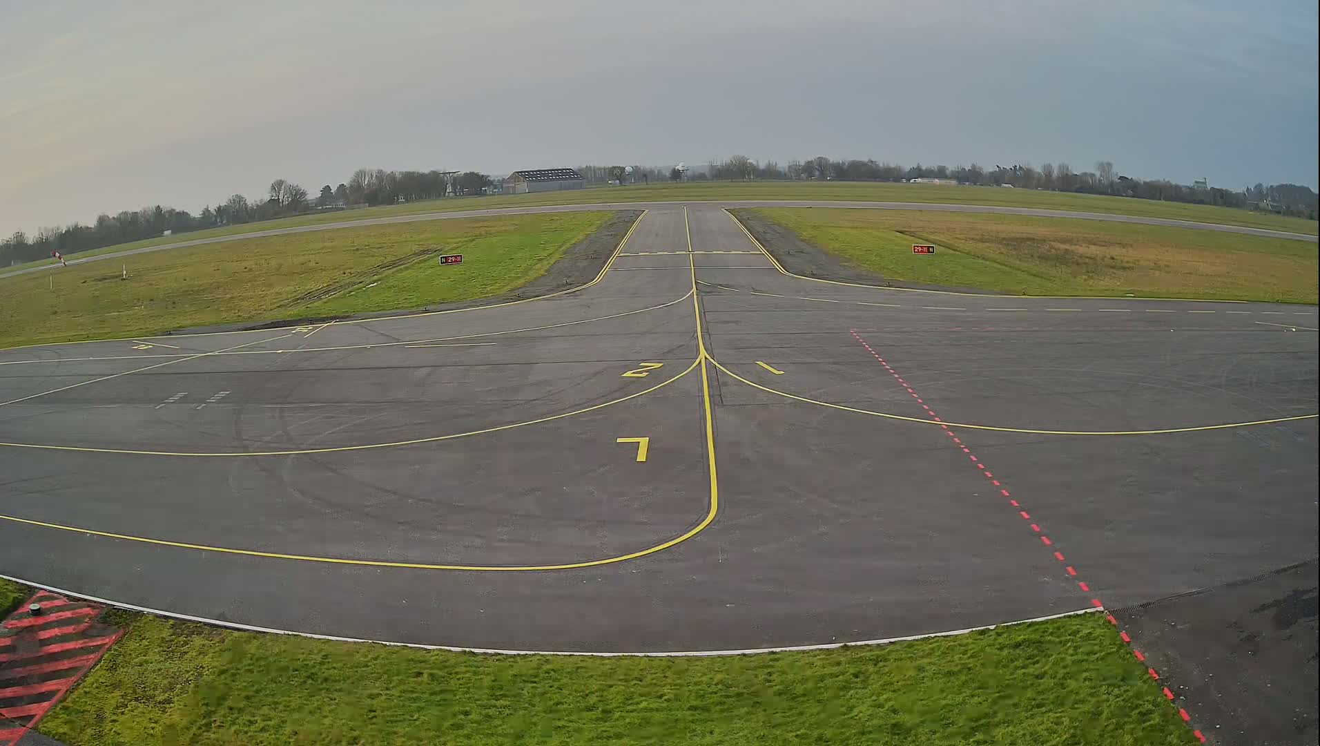 An empty tarmac airport runway area with yellow and red markings is visible under an overcast sky.