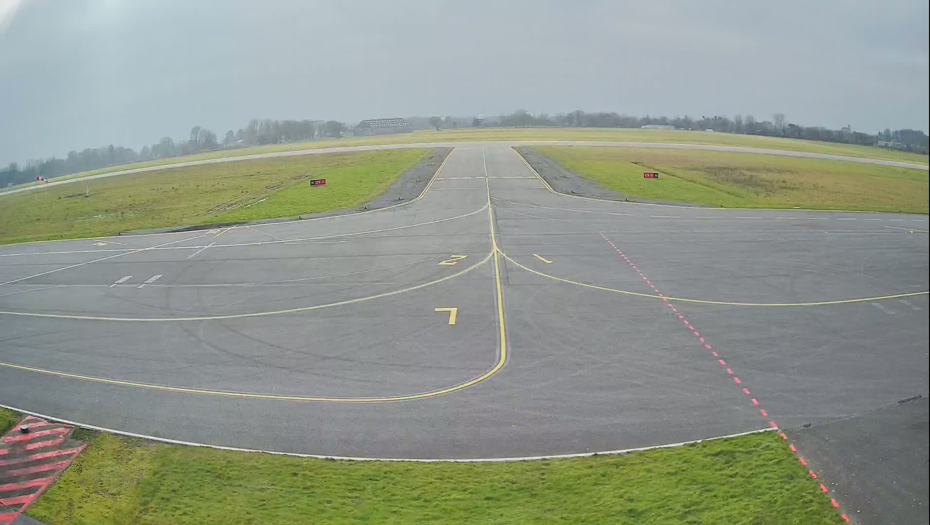 An overcast day at an airfield shows a paved tarmac area with painted lines and numbers, leading to a grassy runway.