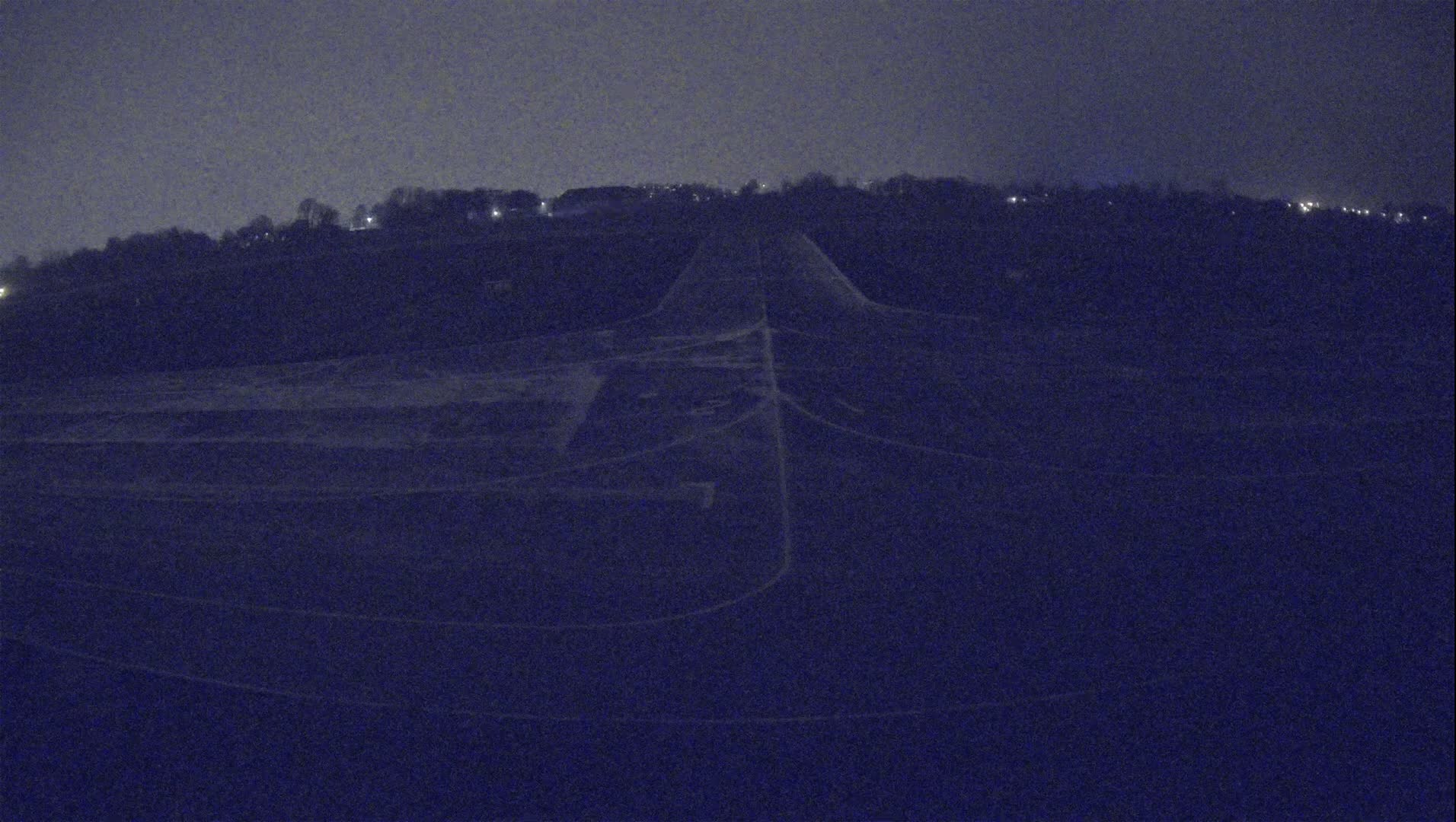 A dark, nighttime view shows a long, paved road or runway extending upward toward a treeline under a dusky sky.
