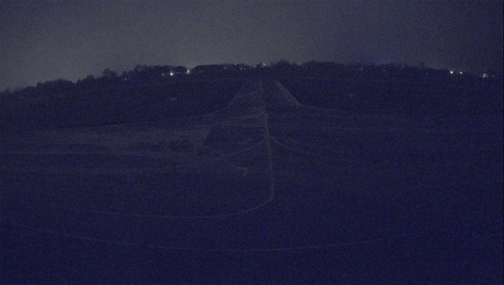 A dark, nighttime view of a long, pale road leading towards a distant hill with faintly visible lights under a dusky sky.