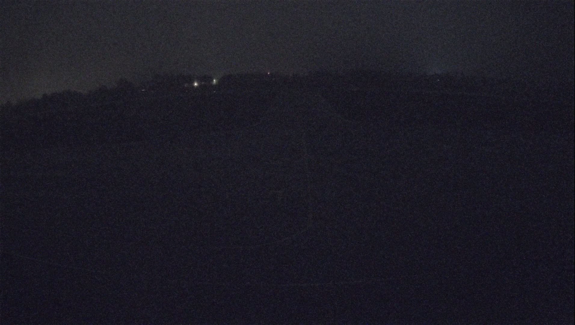 A dark, nighttime landscape shows a distant line of trees and faint lights under a seemingly clear sky.