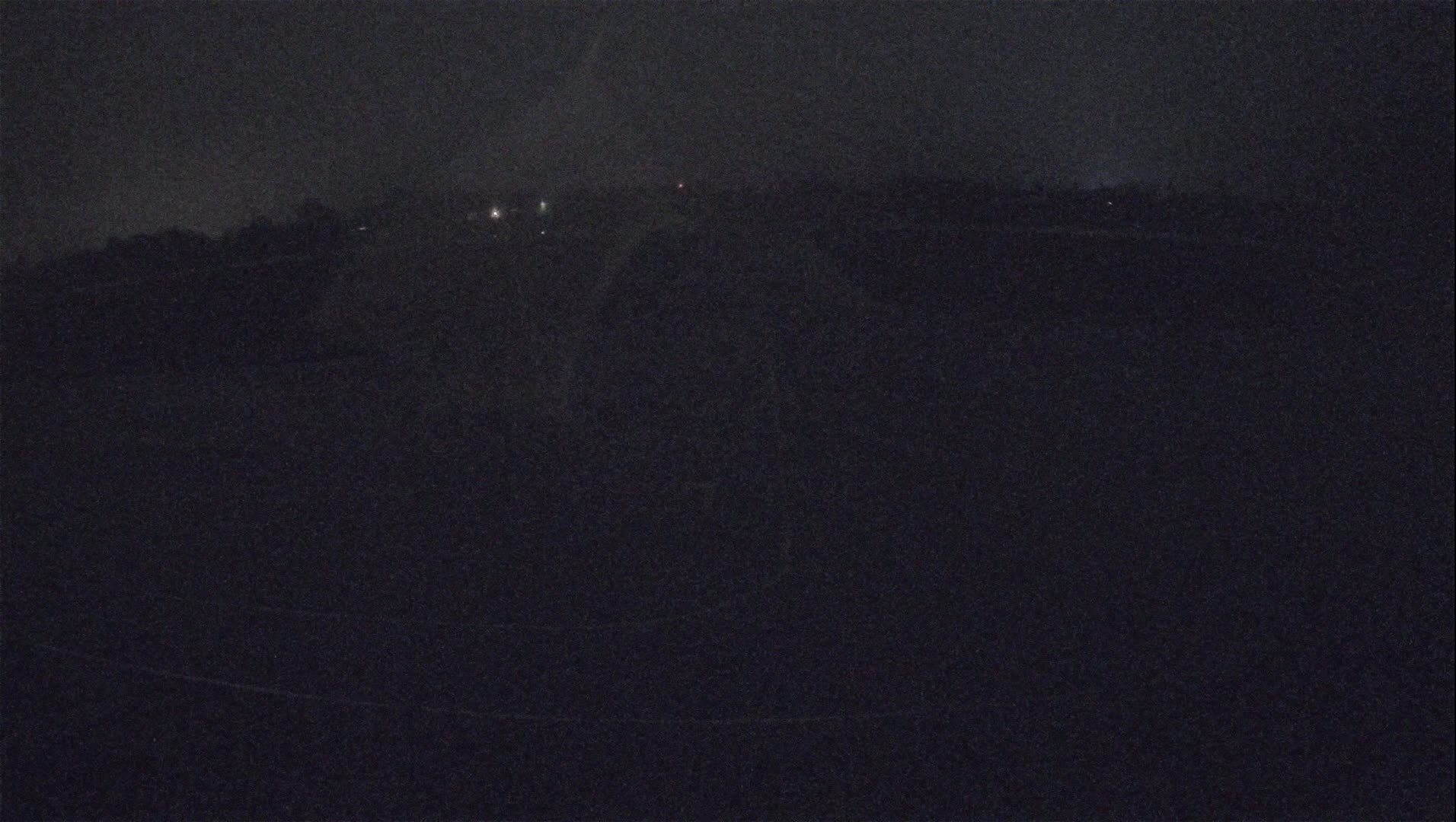 A dark, nighttime scene shows a flat expanse of land with a few distant lights under a hazy sky.