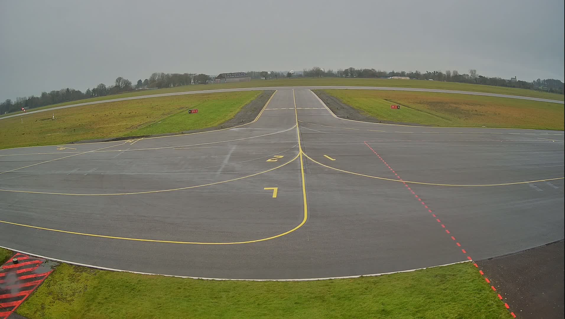 A wet airfield tarmac with yellow and red markings extends toward a runway under an overcast sky.
