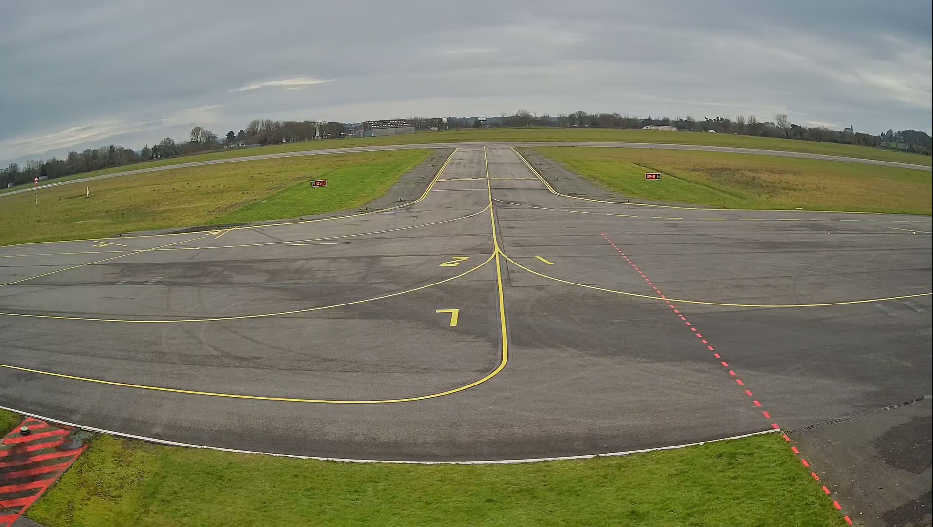 An empty airport tarmac with yellow and red painted lines is shown under an overcast sky.