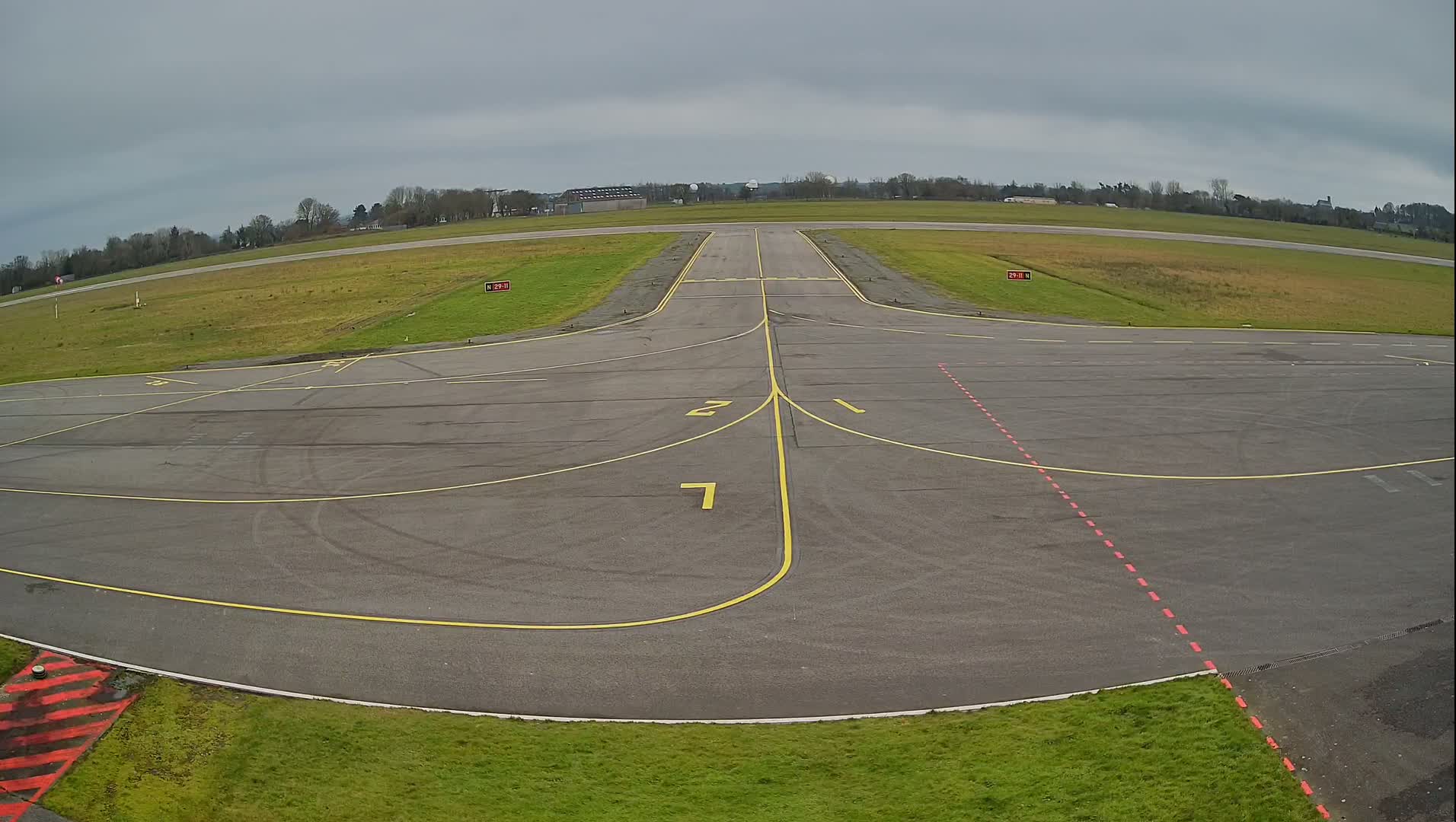 An overcast day shows an airfield's tarmac with yellow and red markings, leading to a runway that extends to a grassy field bordered by trees.