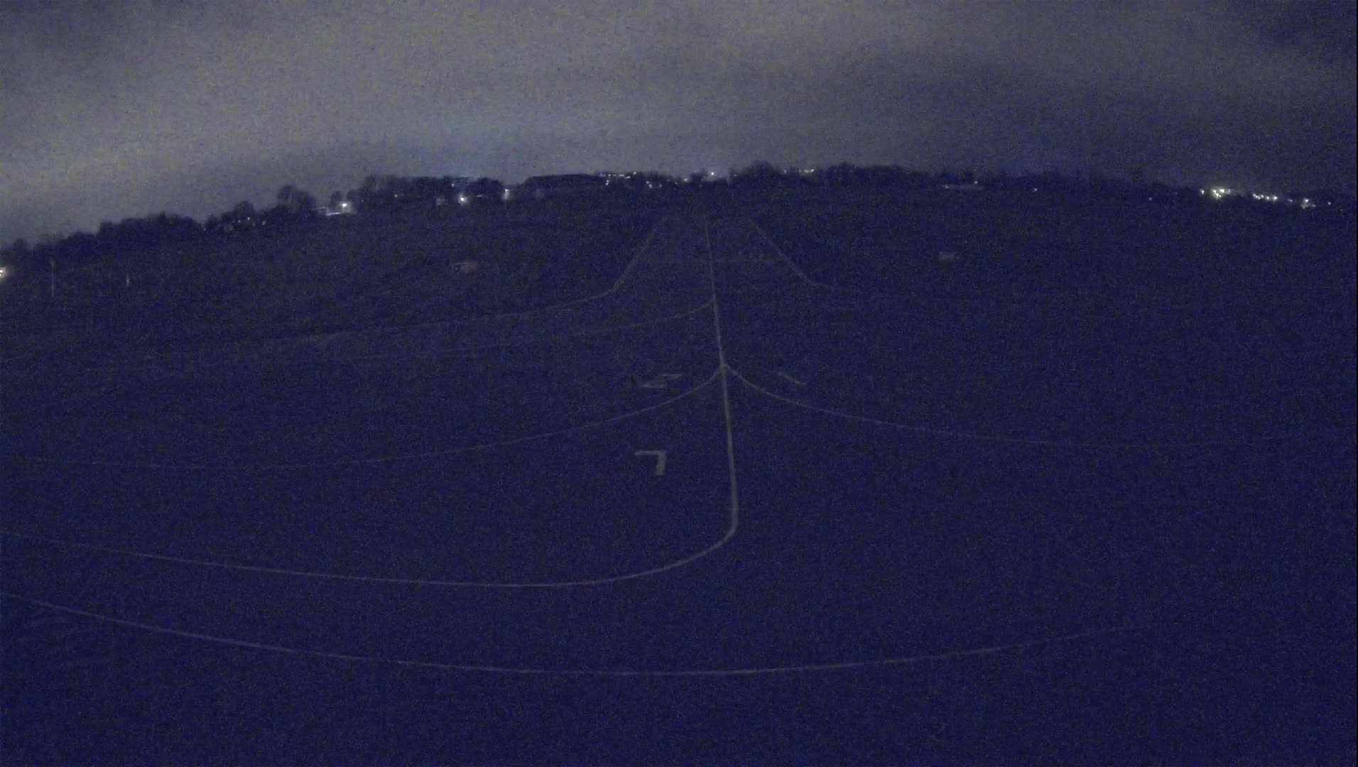 A dark, nighttime view of a large, flat field with faint markings, possibly a sports field, under a cloudy sky.