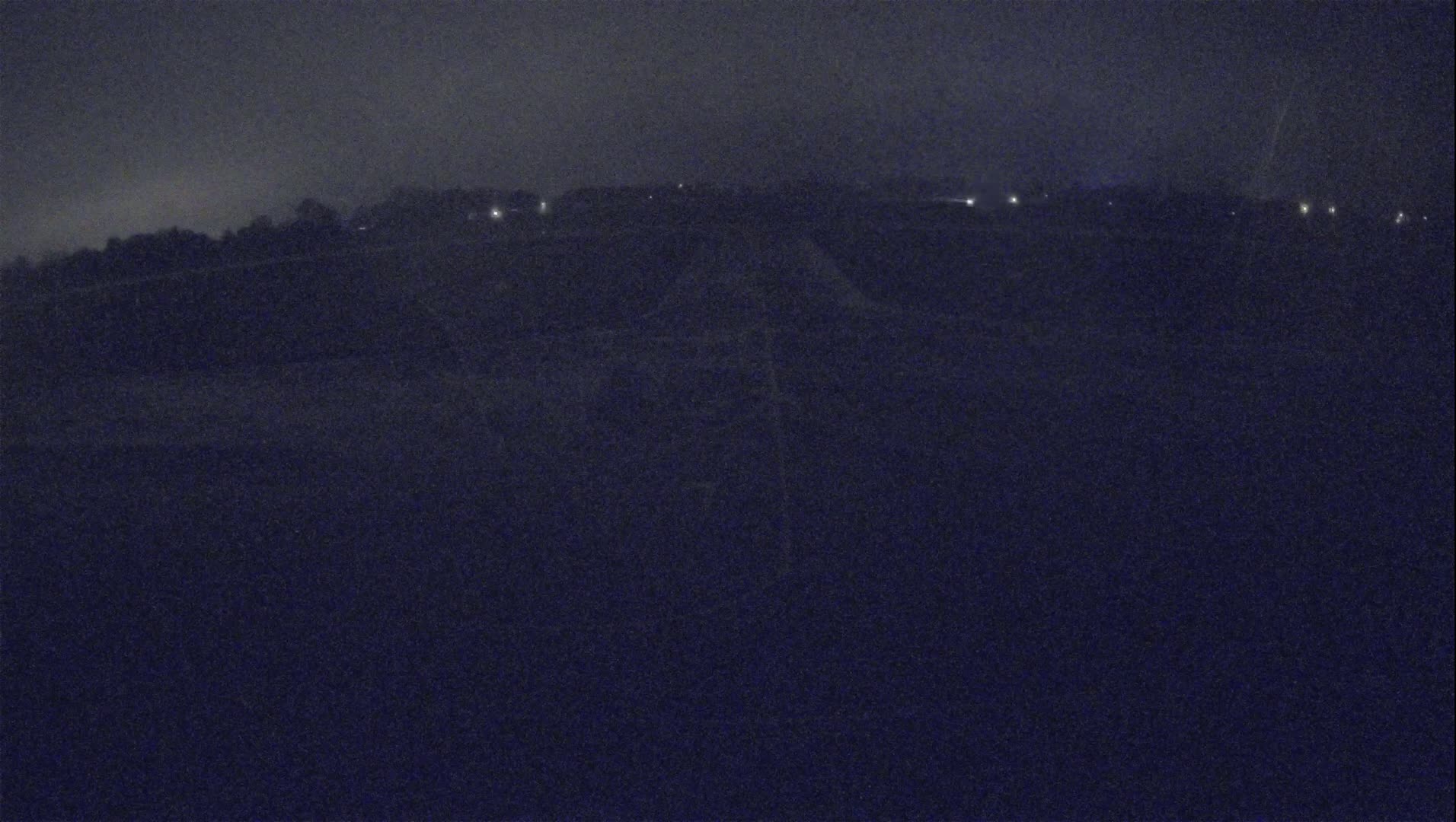 A dark, nighttime landscape with a few distant lights visible under a foggy sky.