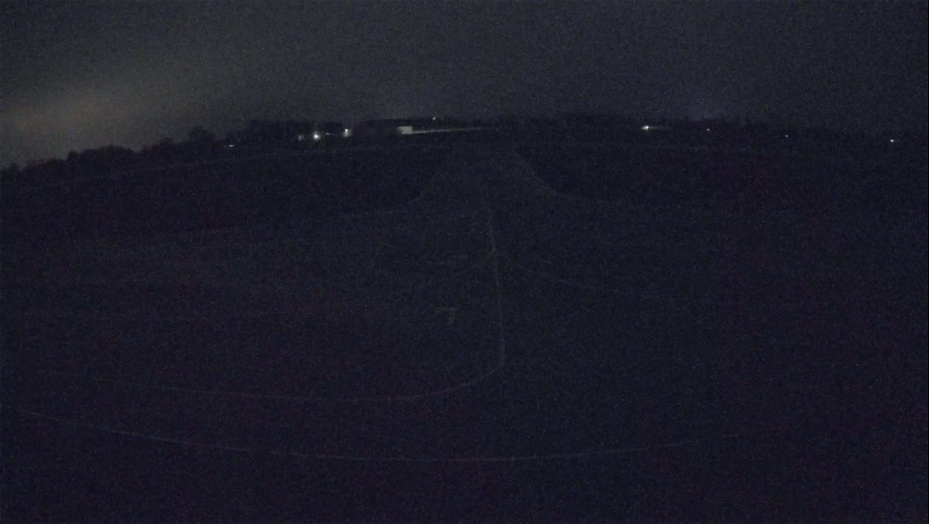 A dark, nighttime view of a flat, empty field with a few faint lights visible in the distance under a cloudy sky.