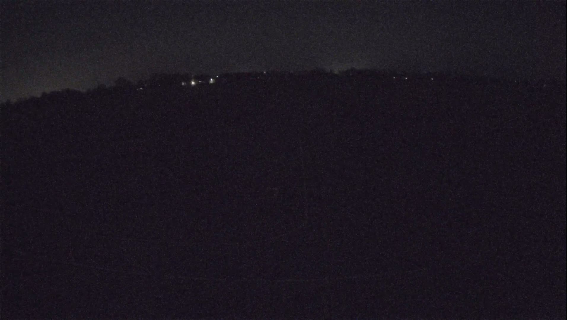 A dark, nighttime landscape shows a distant line of faint lights on a hill under a mostly clear sky.