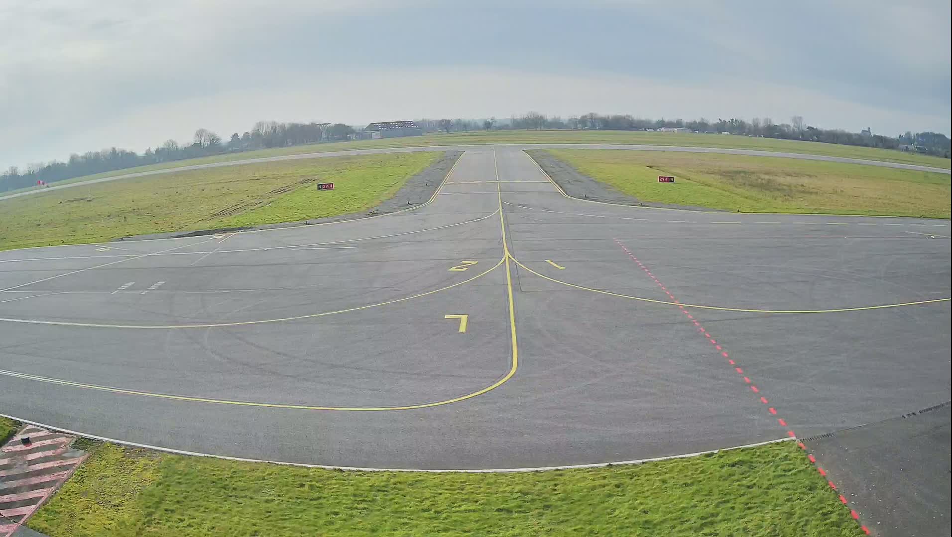 An empty airport tarmac with yellow and red markings is surrounded by short green grass under a cloudy sky.