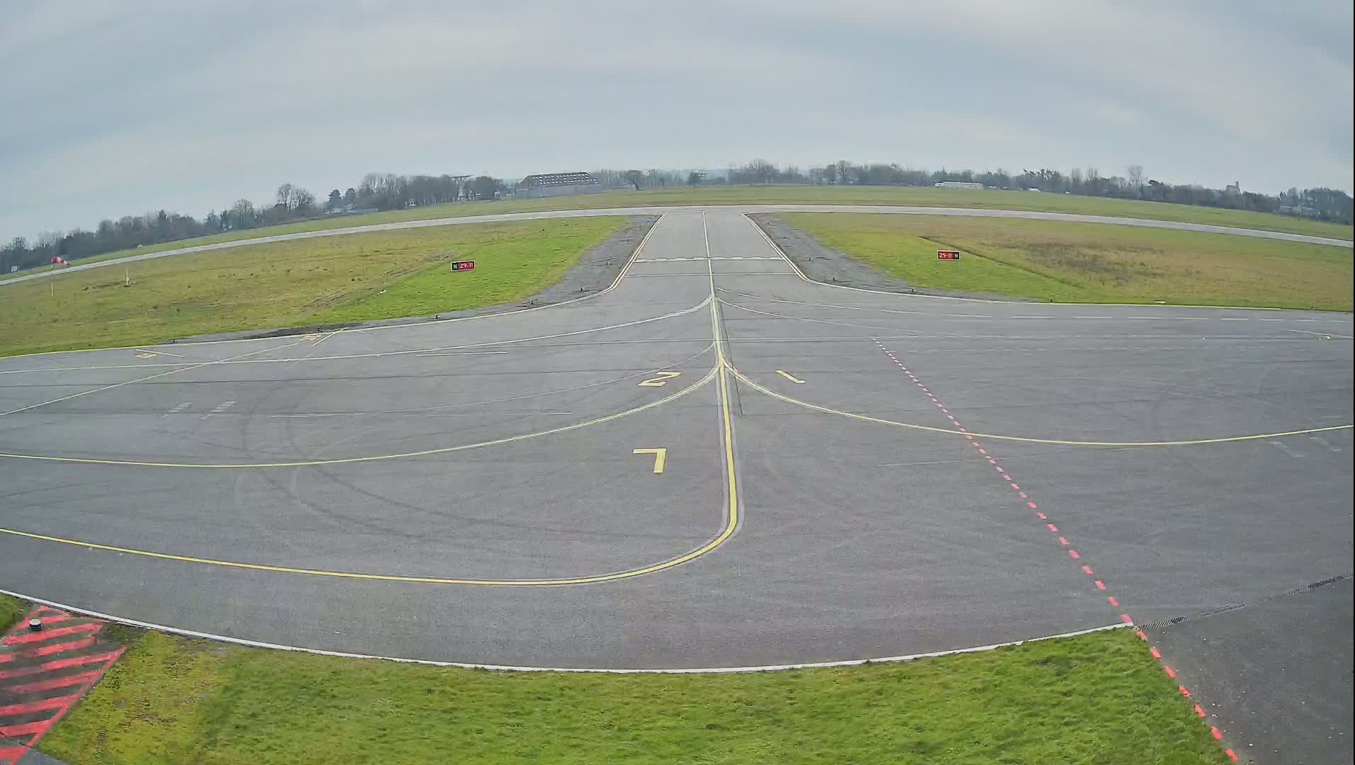 An empty airfield with a runway and taxiway markings is visible under an overcast sky.