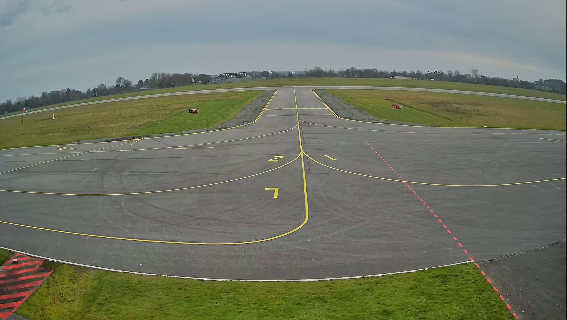 An overcast sky covers a tarmac airport runway with yellow and red markings, surrounded by short green grass.