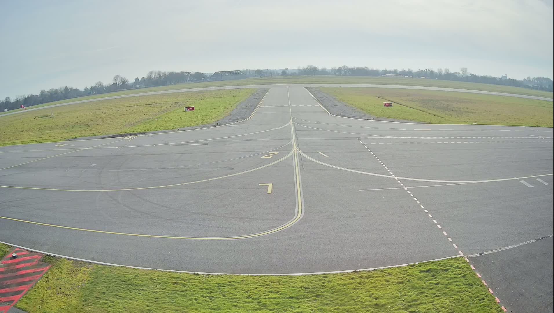 A paved airport taxiway with yellow lines and numbers, surrounded by short green grass under an overcast sky.