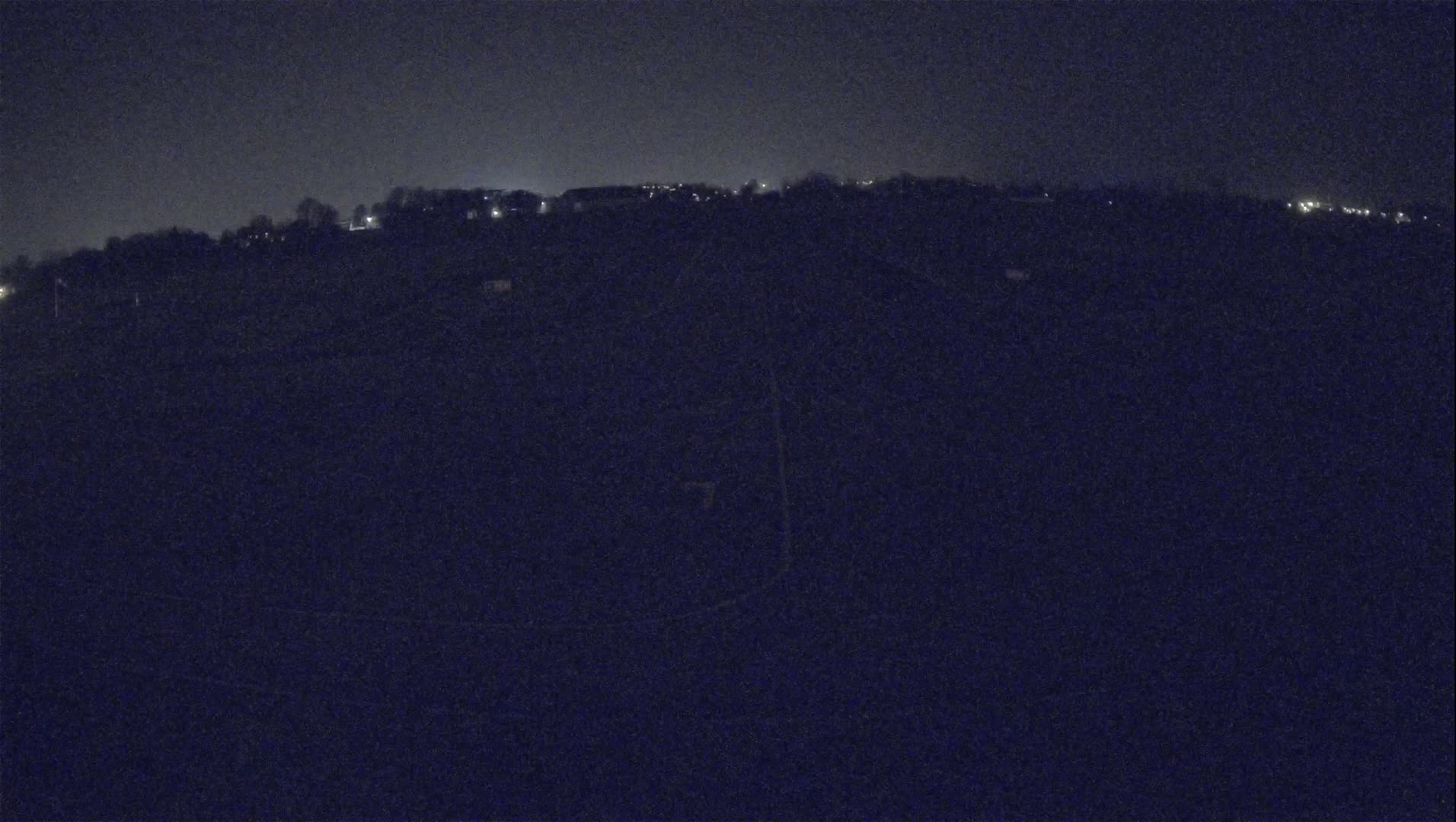 A dark, nighttime landscape shows a distant line of dimly lit buildings on a hill under a mostly clear sky.