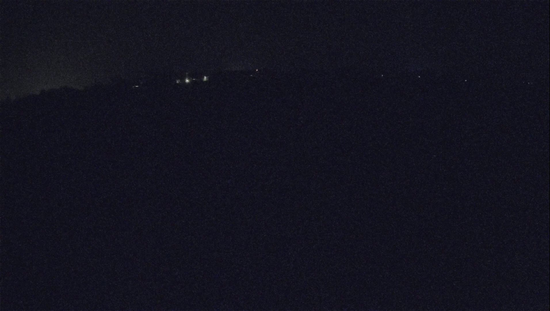 A dark, nighttime landscape shows a distant line of faint lights on a hill.