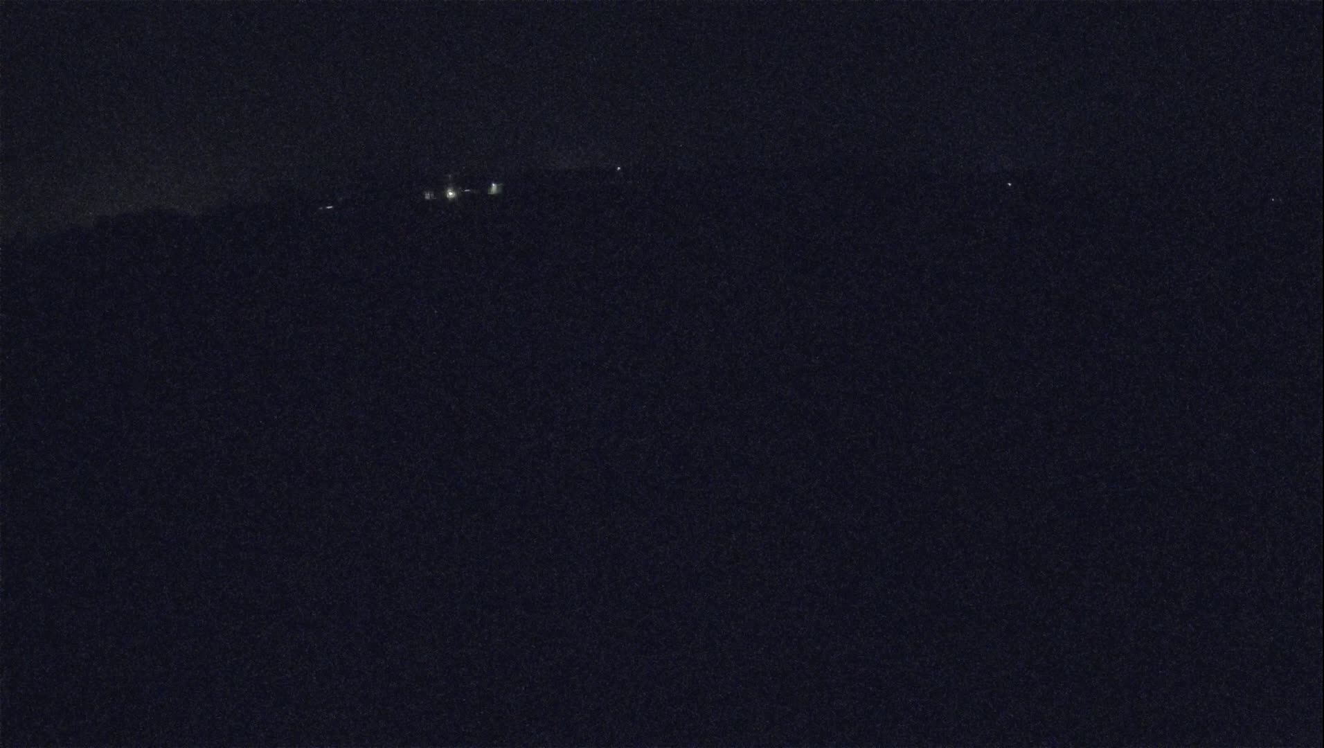 A dark, nighttime landscape shows a few distant lights on a hill under a clear sky.