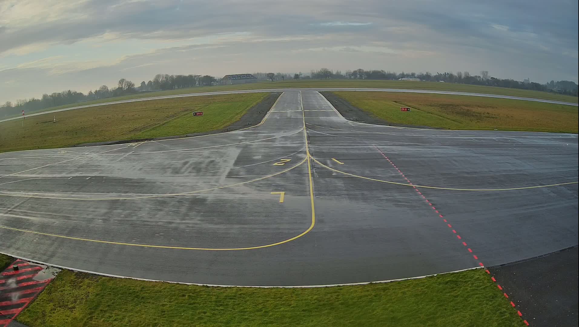 A wet airport tarmac with runway markings is visible under a cloudy sky.