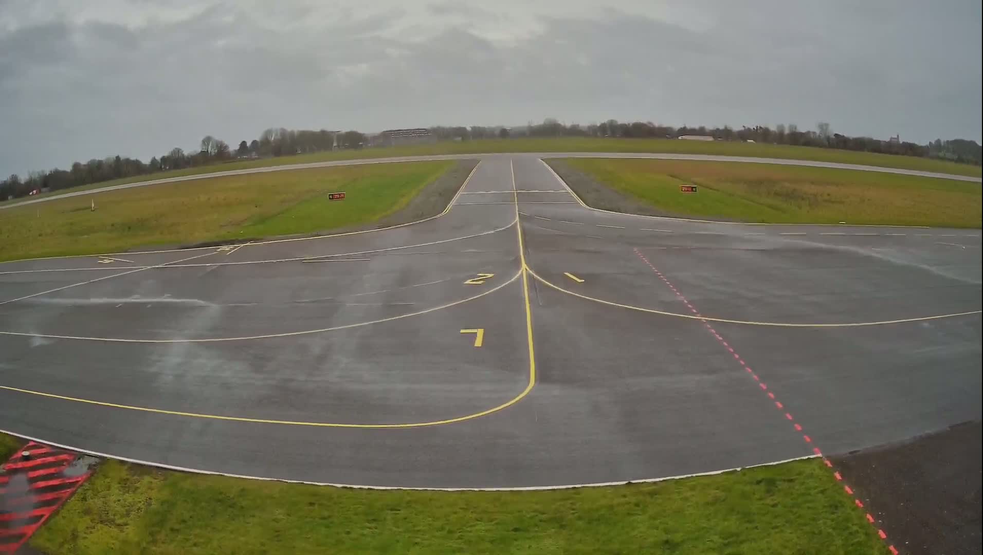 A wet, paved airport runway with yellow and red markings is visible under a cloudy sky.