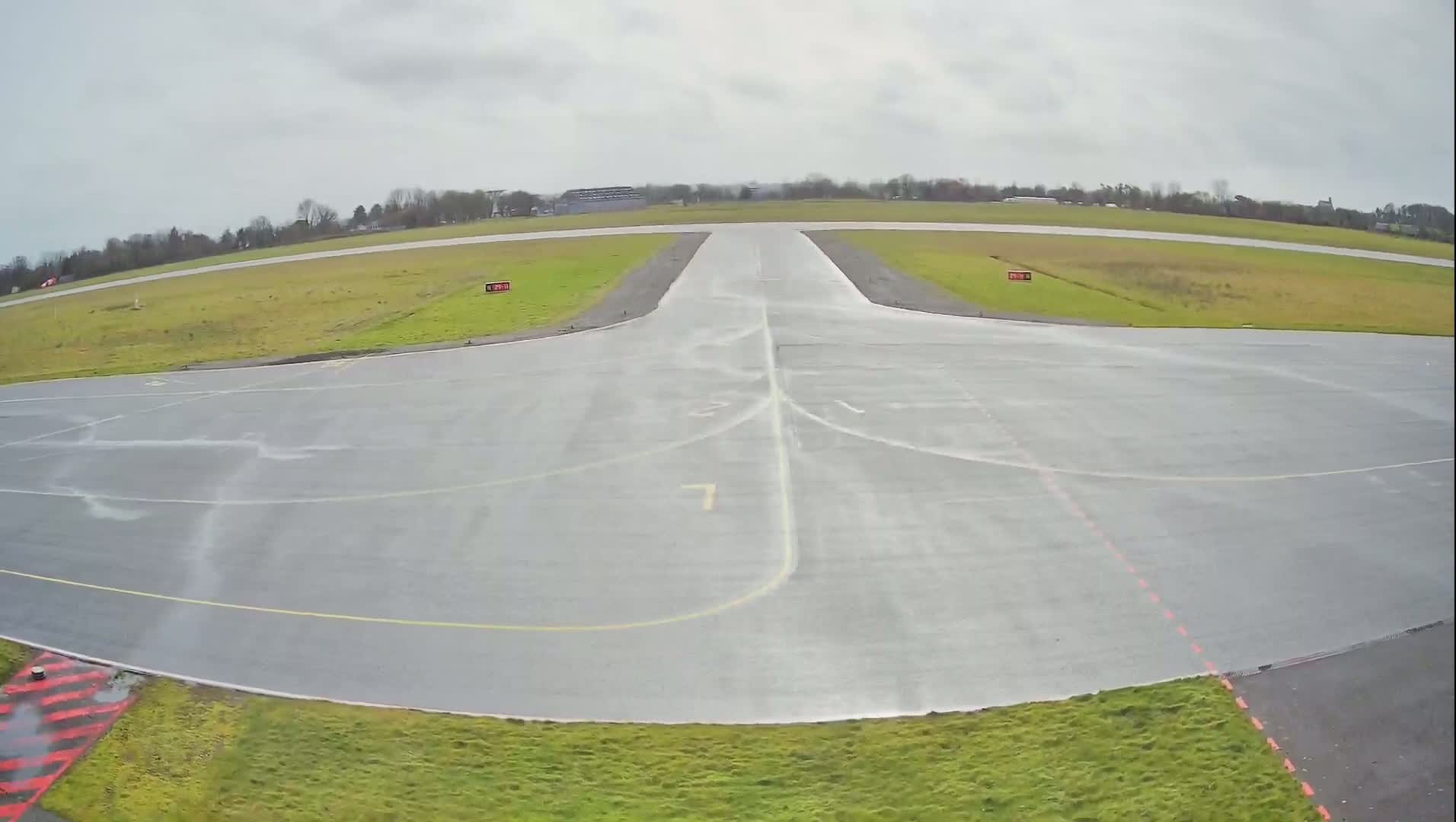 A wet airport tarmac with a runway and surrounding grassy areas under an overcast sky.