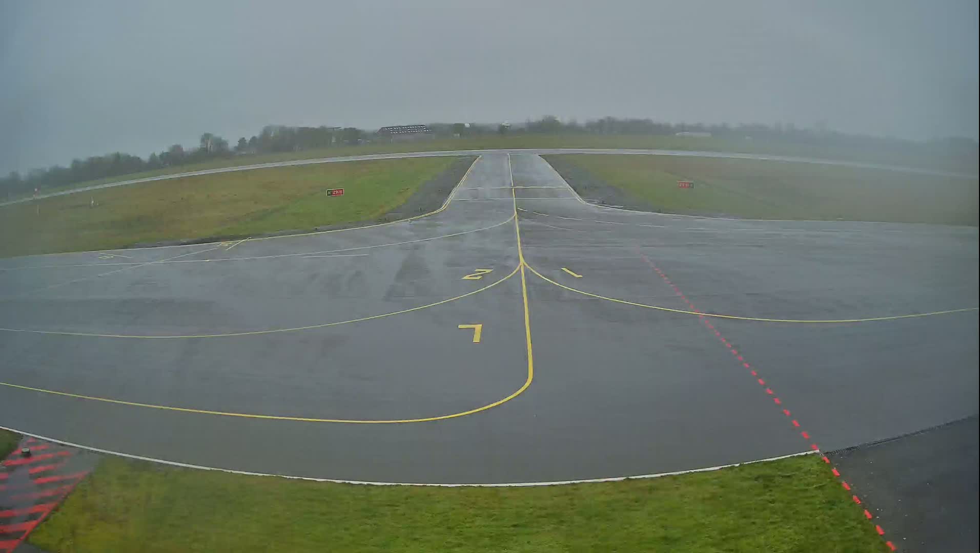 A wet, gray tarmac airport runway is visible, with surrounding grassy areas under an overcast sky.