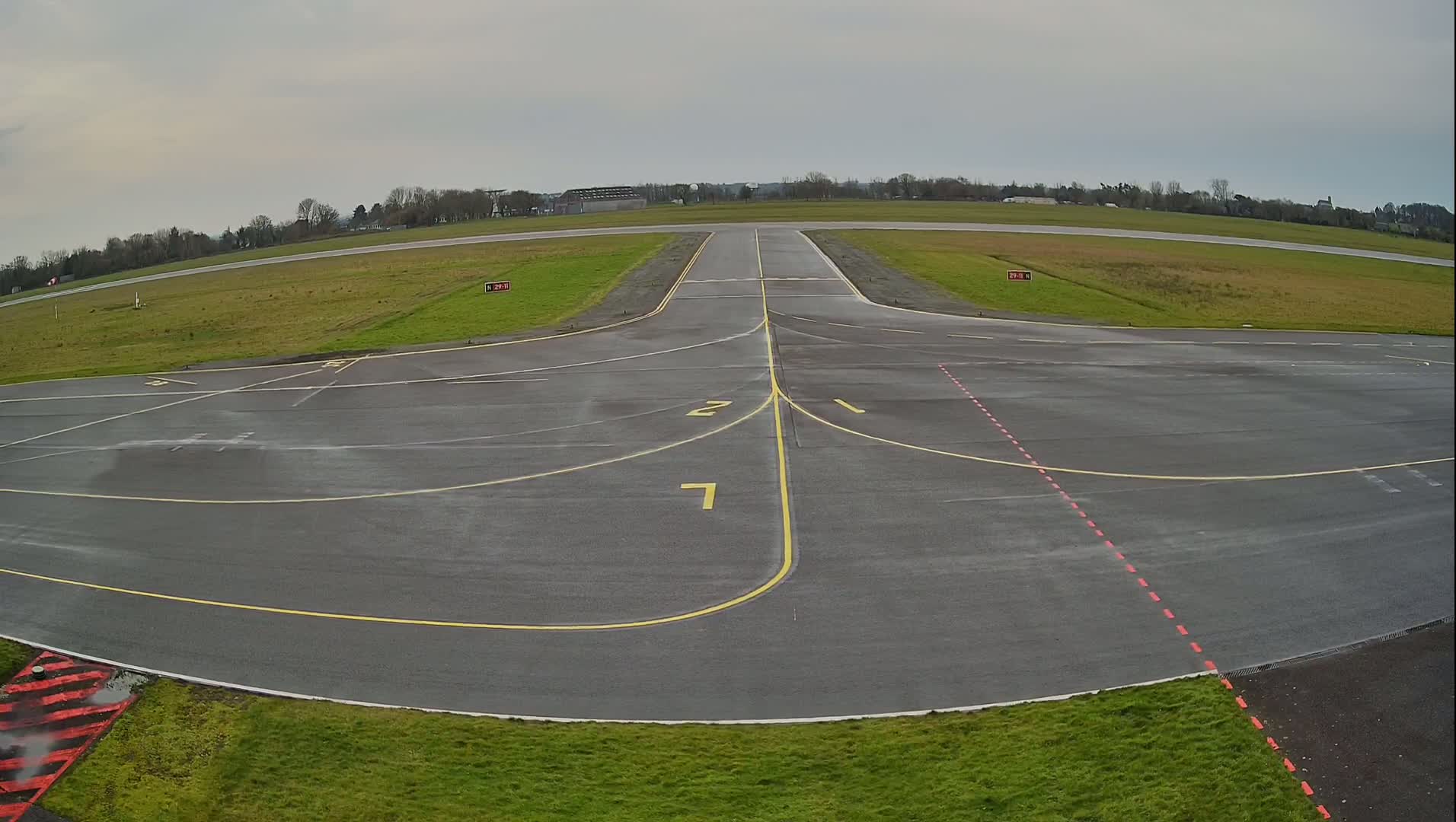 A wet airport tarmac shows runway markings and taxiways under an overcast sky.