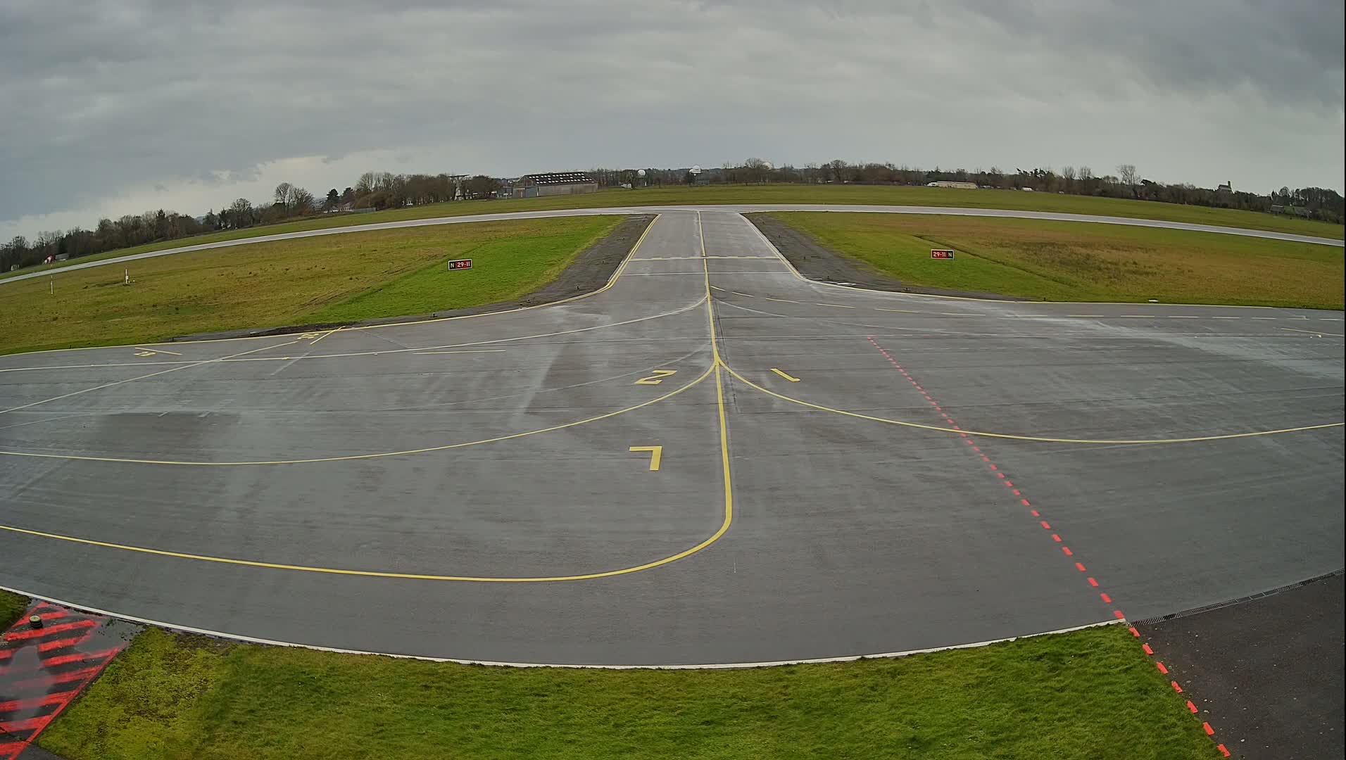 A wet, gray airport tarmac with yellow and red markings under an overcast sky.